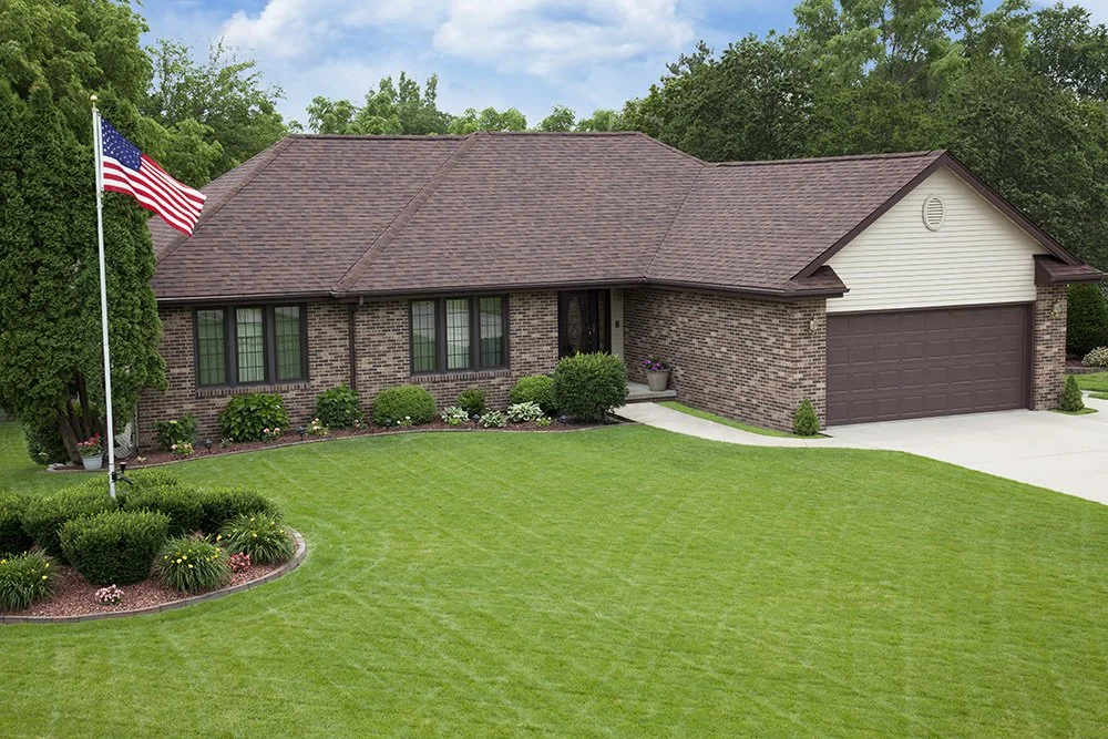 Front view of a single-story brick house with a two-car garage, manicured lawn, American flag on a pole, and trees in the background.