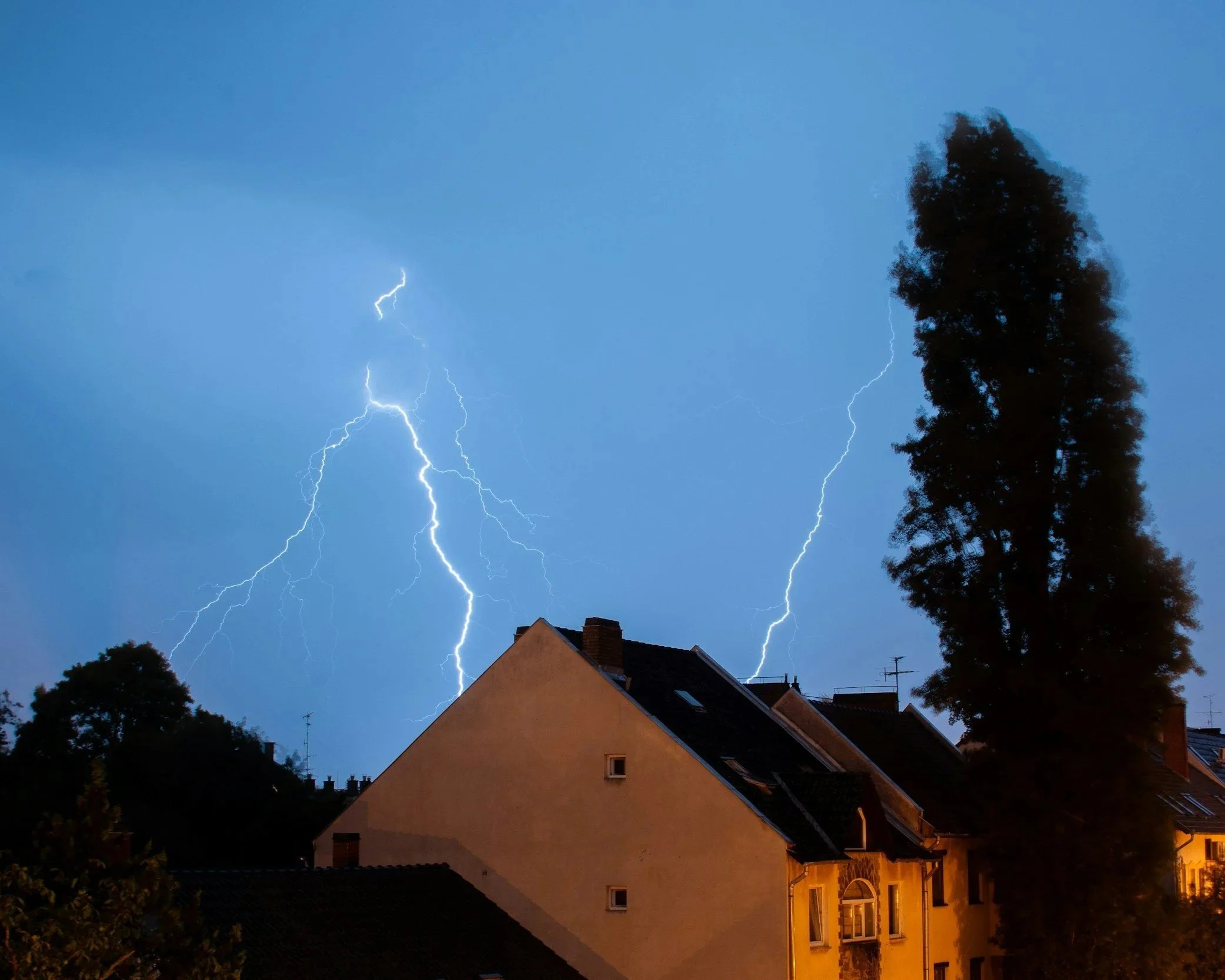 Lightning strikes over houses and trees at night in a residential neighborhood.