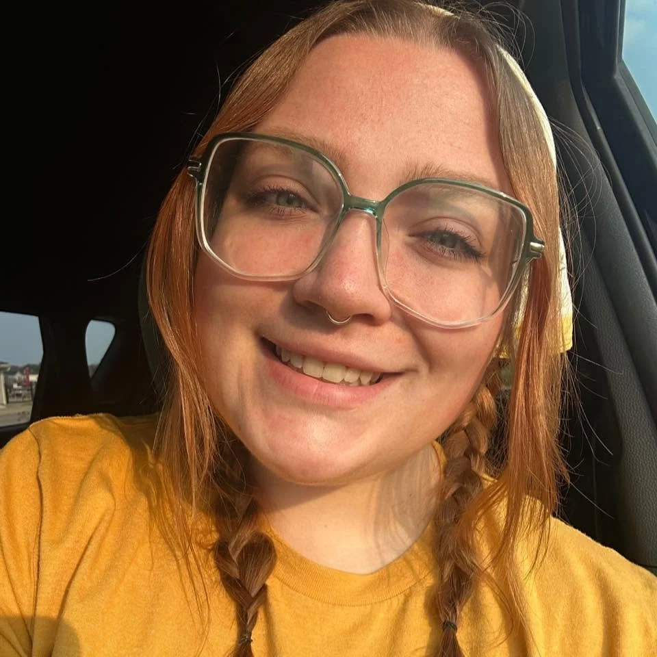 A young woman with red hair and glasses smiling in a car.