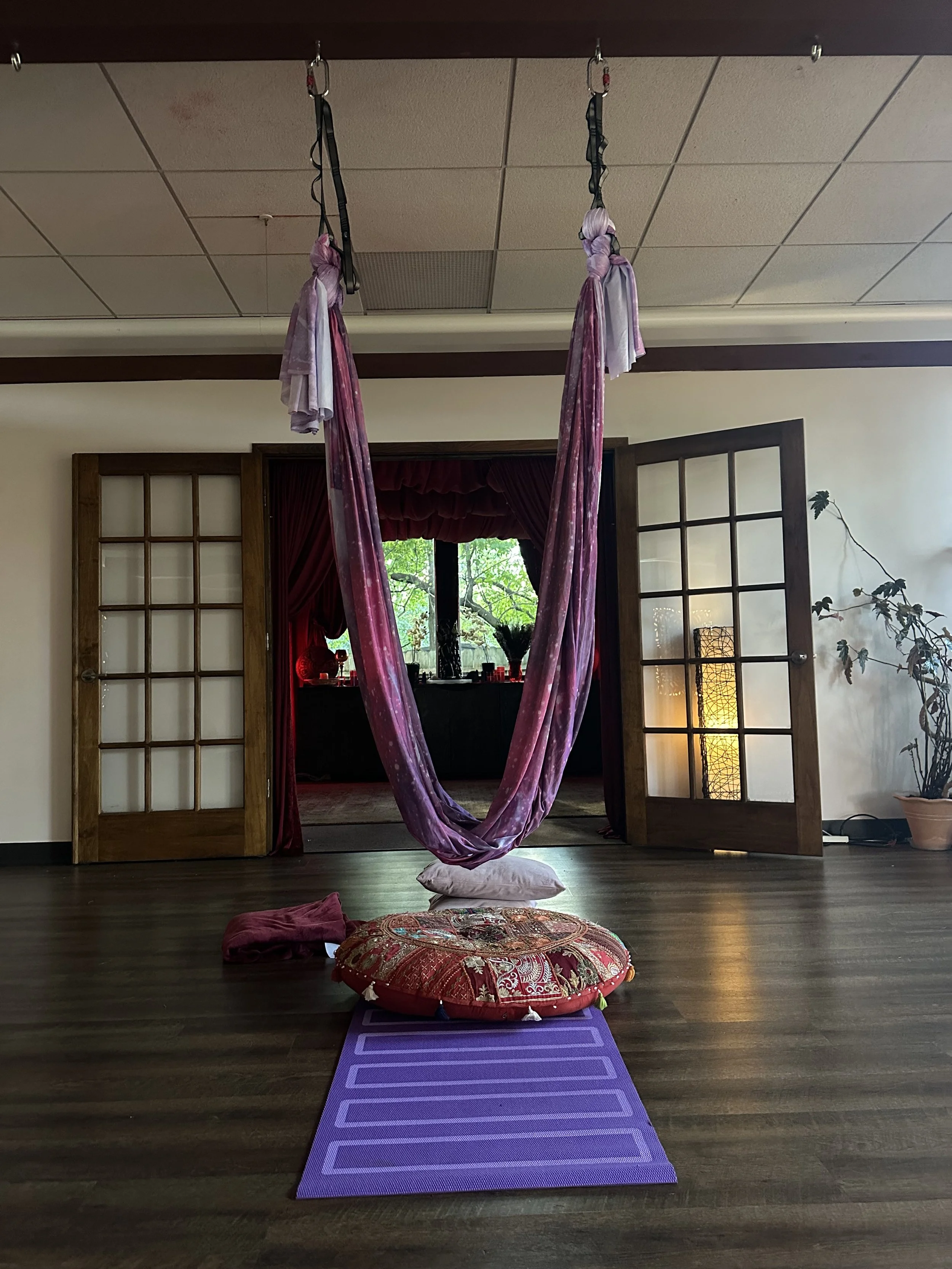 Aerial yoga setup with purple yoga mat, round decorative cushion, hanging pink silk hammock, pillows, and a window with green trees outside.