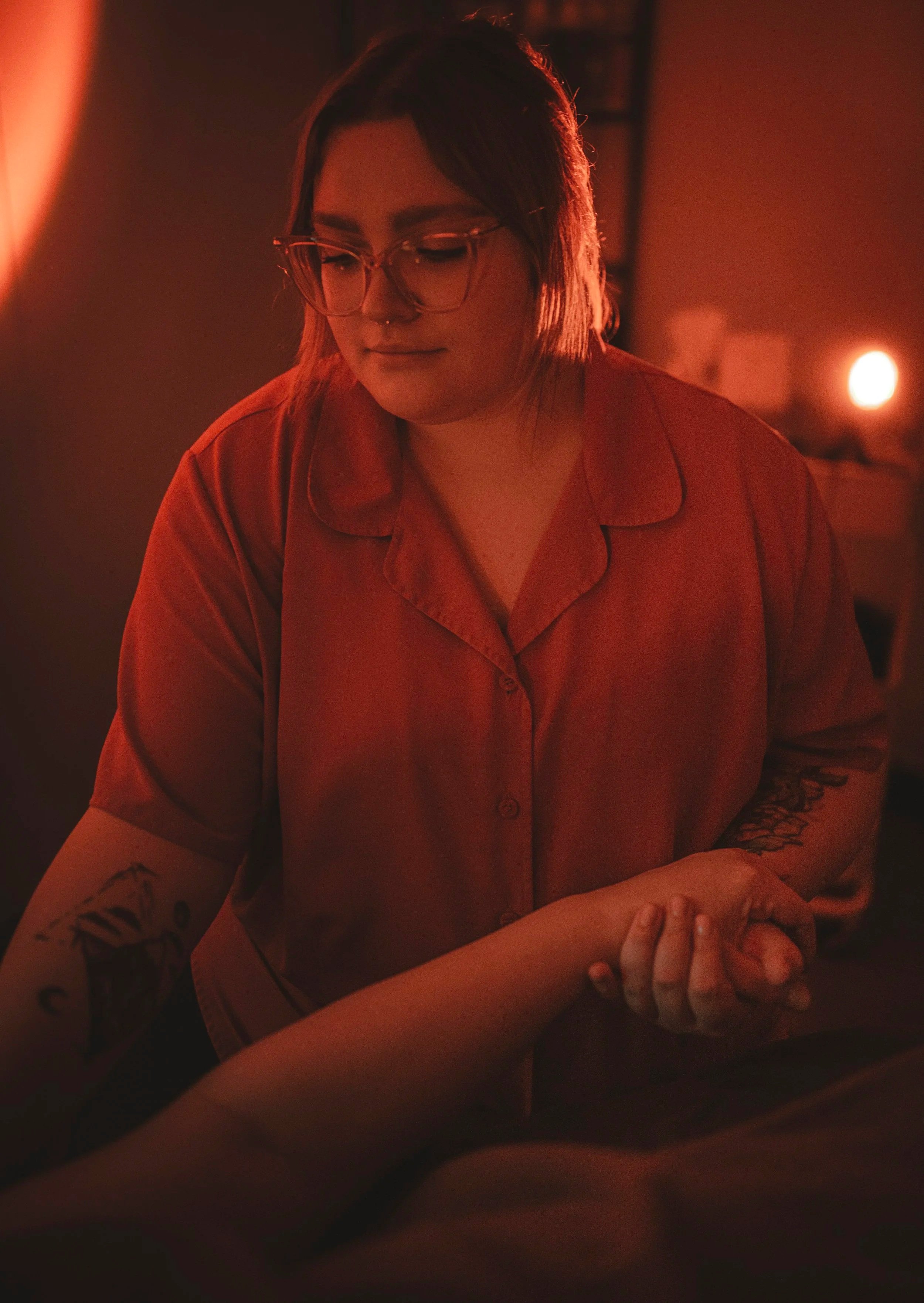 A woman with glasses and tattoos on her arm, wearing an orange shirt, sitting in a dimly lit environment with red lighting.