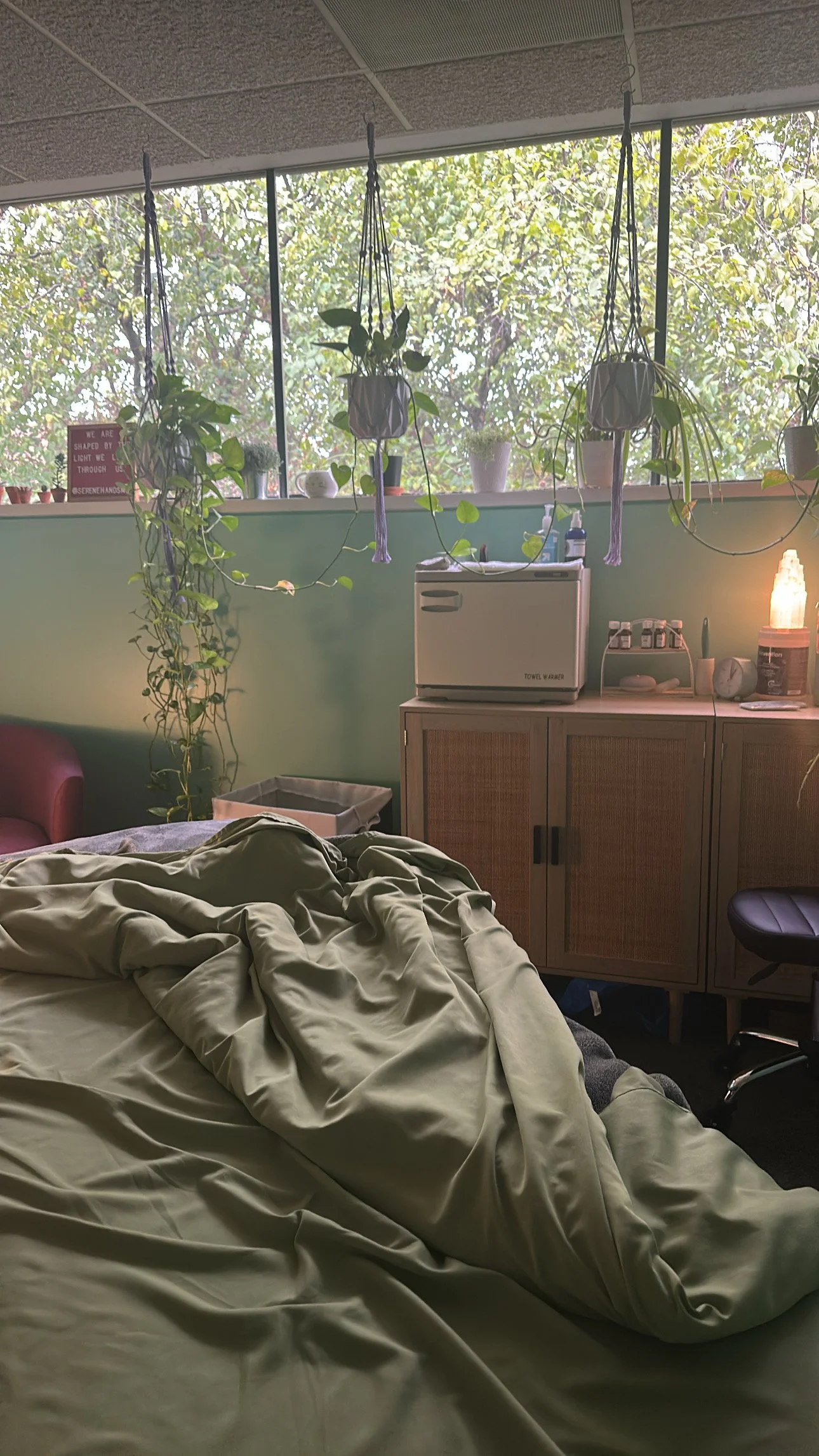 A bedroom with an unmade bed, green bedding, and a window with plants on the windowsill and hanging from the ceiling. There is a small fridge, bottles, and a salt lamp on a wooden cabinet. The room has a relaxed, cozy atmosphere.