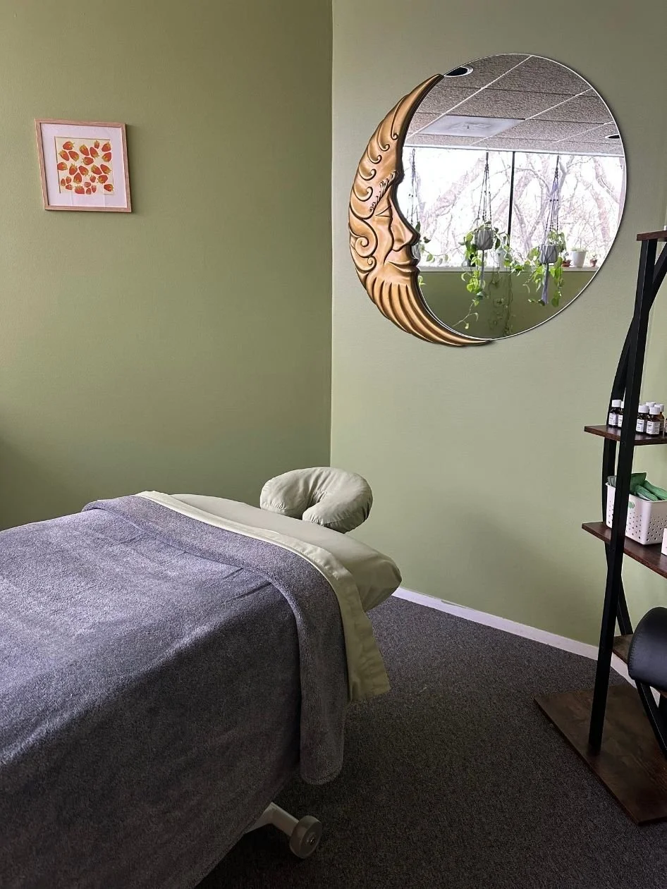 Massage room with a massage table covered in gray blanket, a face cradle, a round mirror with a crescent moon face frame, potted plants on windowsill, and wall art of leaves.
