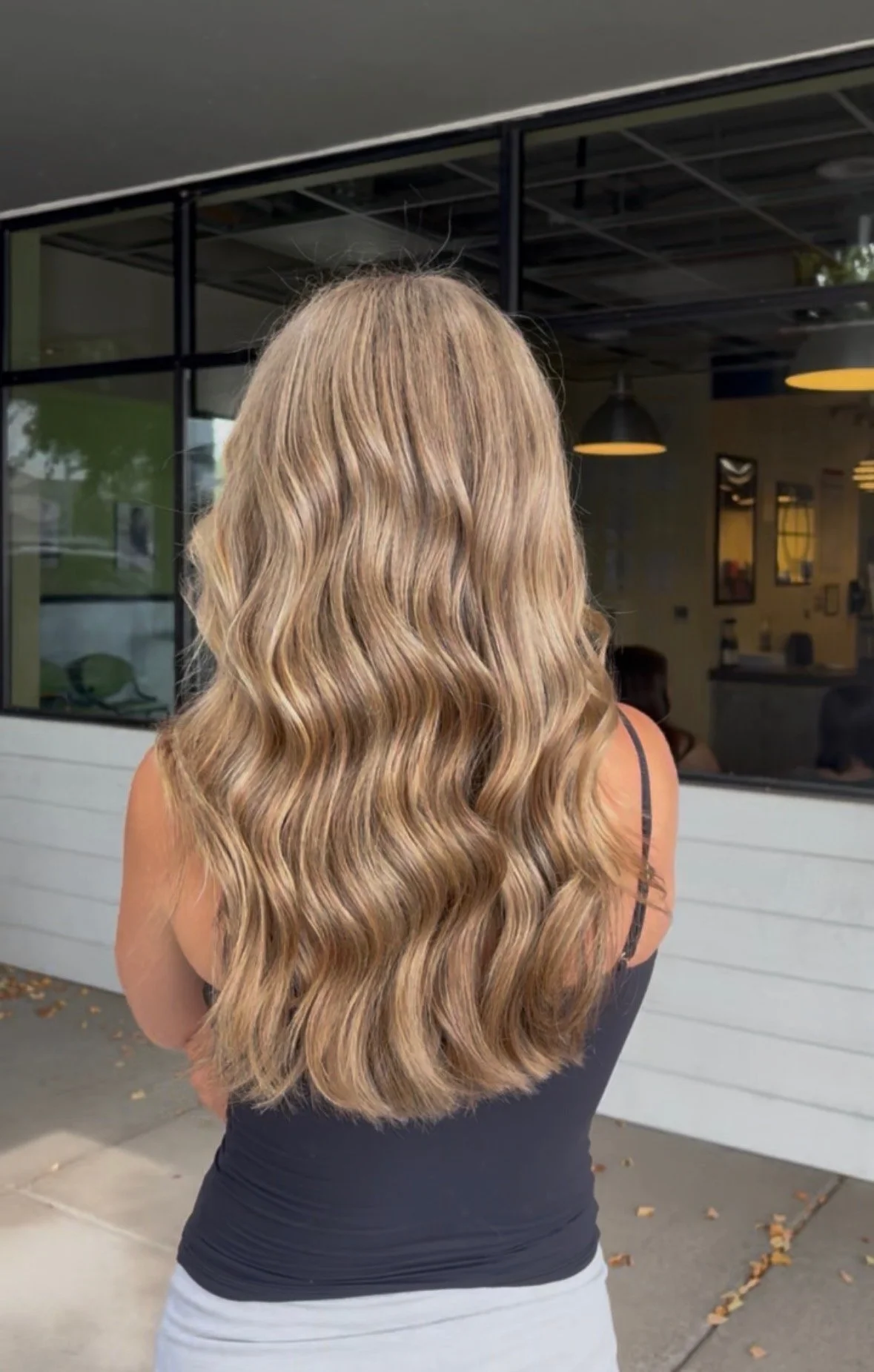 Back view of woman with long, wavy blonde hair. She is outdoors, wearing a black tank top and light gray pants, looking toward a building with glass windows and hanging lights.