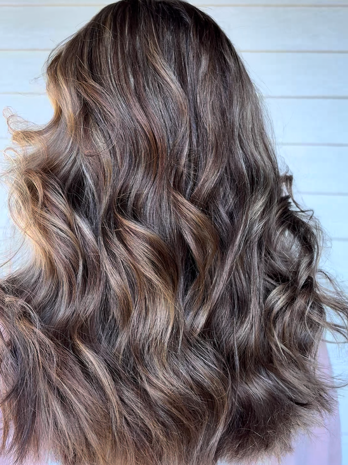 Close-up of wavy, multi-colored hair with shades of brown, blonde, and ash gray, falling in loose curls against a light blue background.