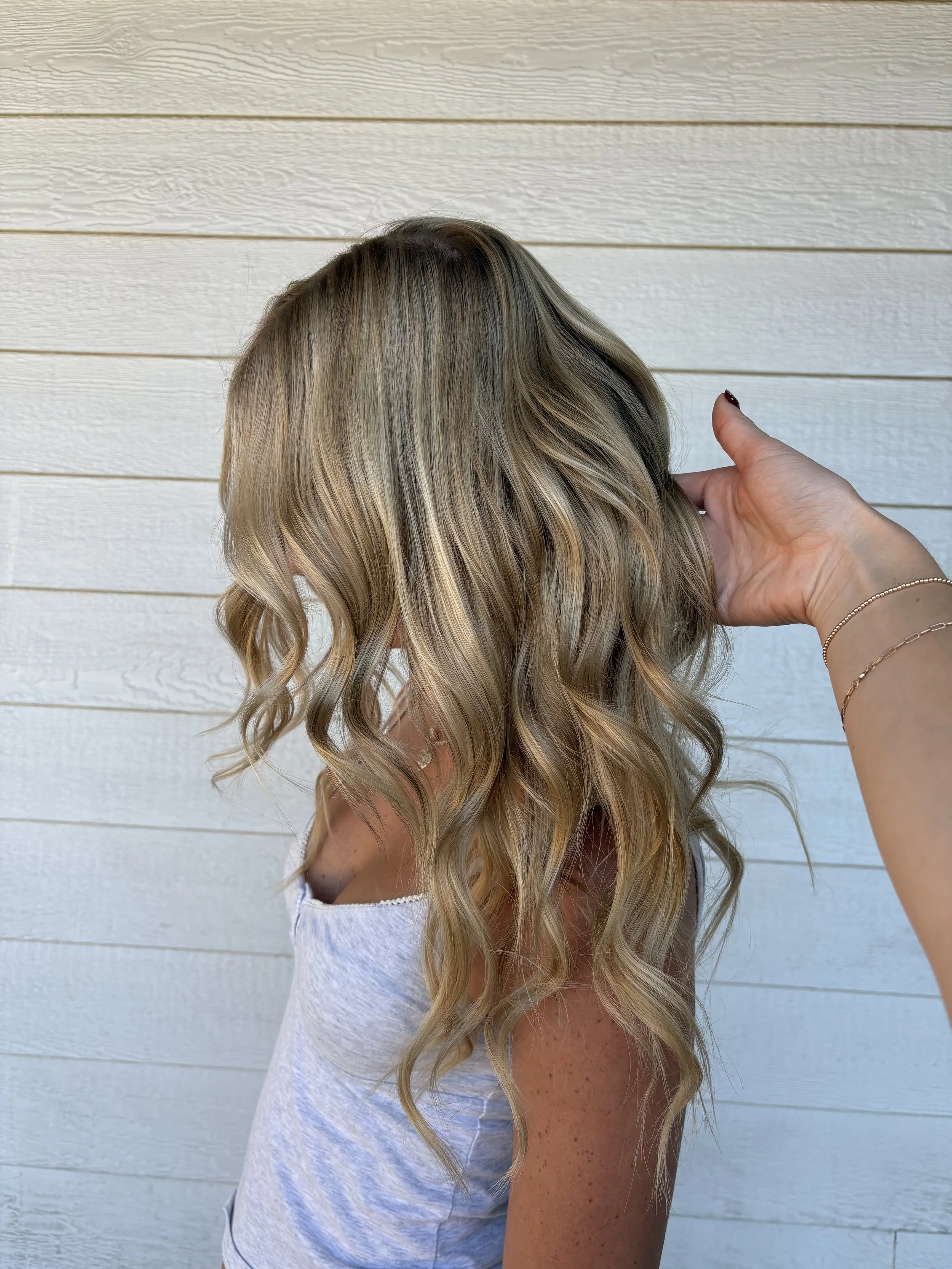 A woman with long, wavy blonde hair standing against a white wooden wall, with her hand touching her hair.