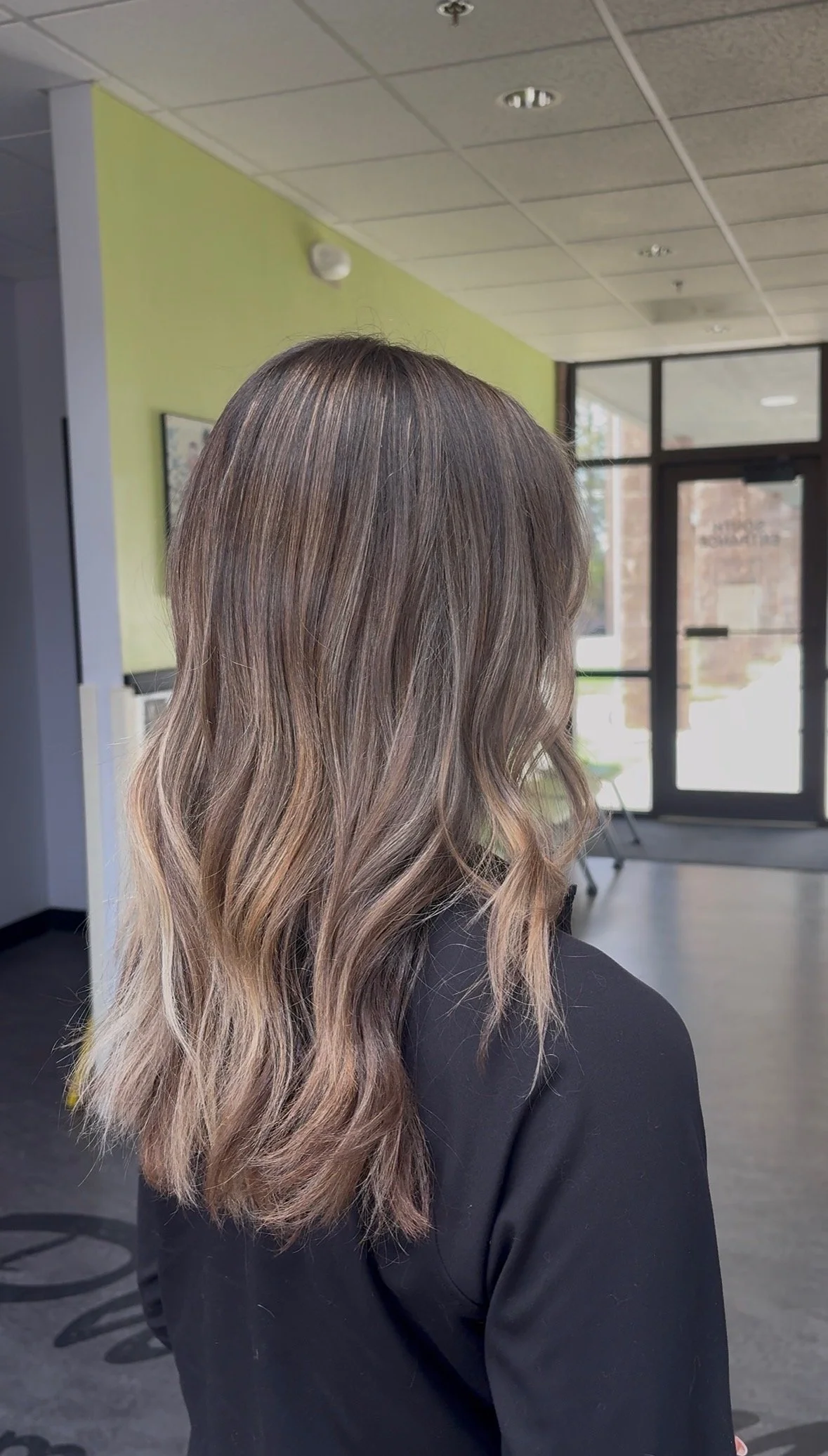 A woman with wavy, shoulder-length hair that is dark brown on top and fades into a blonde ombre at the ends, standing indoors near a light green wall and large glass door.