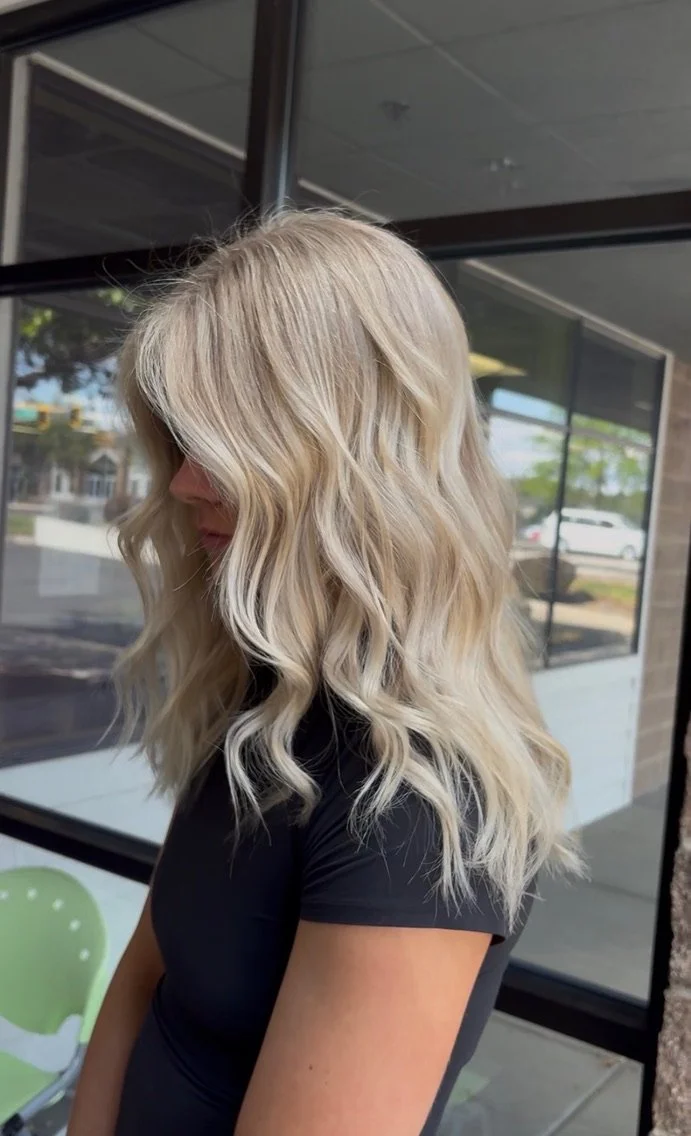 Side view of a woman with long, wavy blonde hair wearing a black shirt, standing outdoors near a building with large windows.