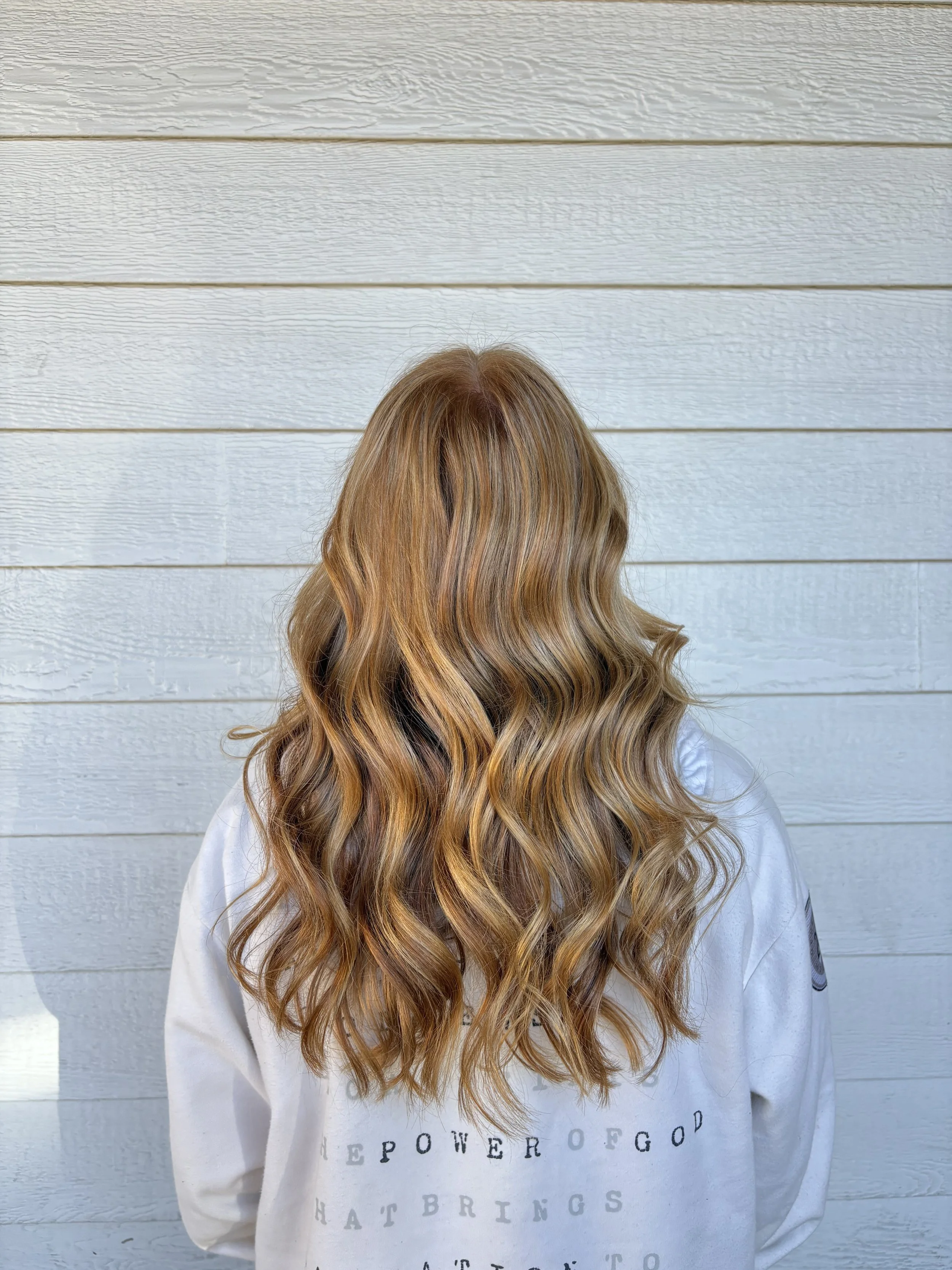 Person with long, wavy, brown hair, facing away from the camera, wearing a white sweatshirt with black text, standing in front of a white wooden wall.