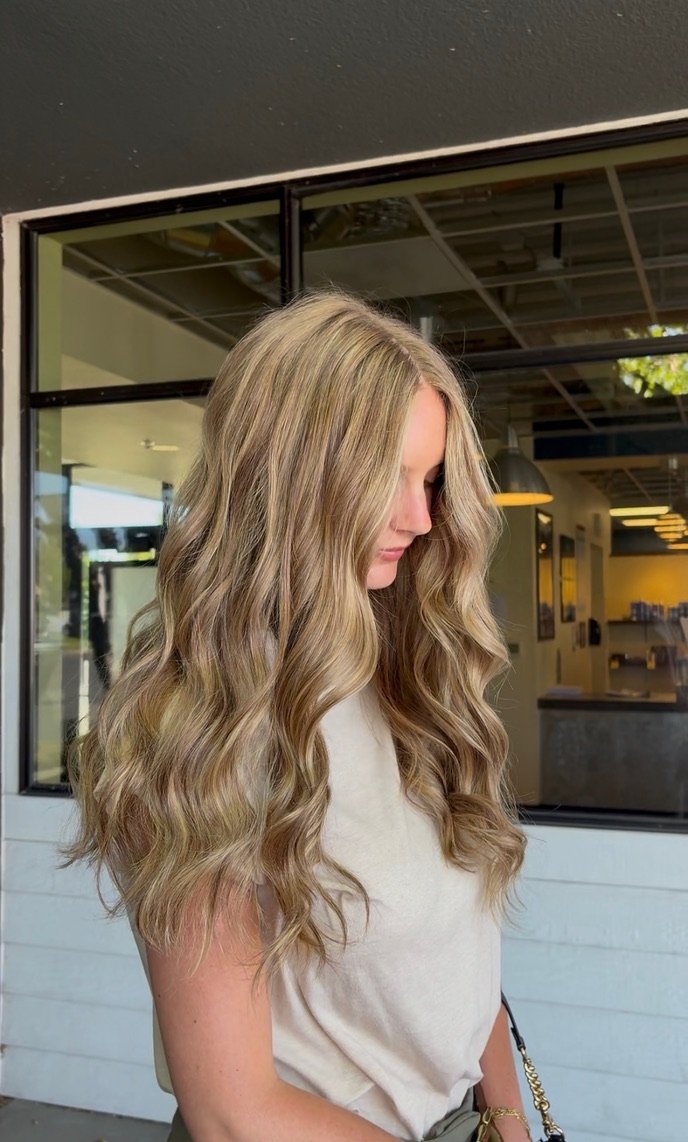 Side profile of a blonde woman with wavy hair standing indoors near a large window, wearing a light-colored top.