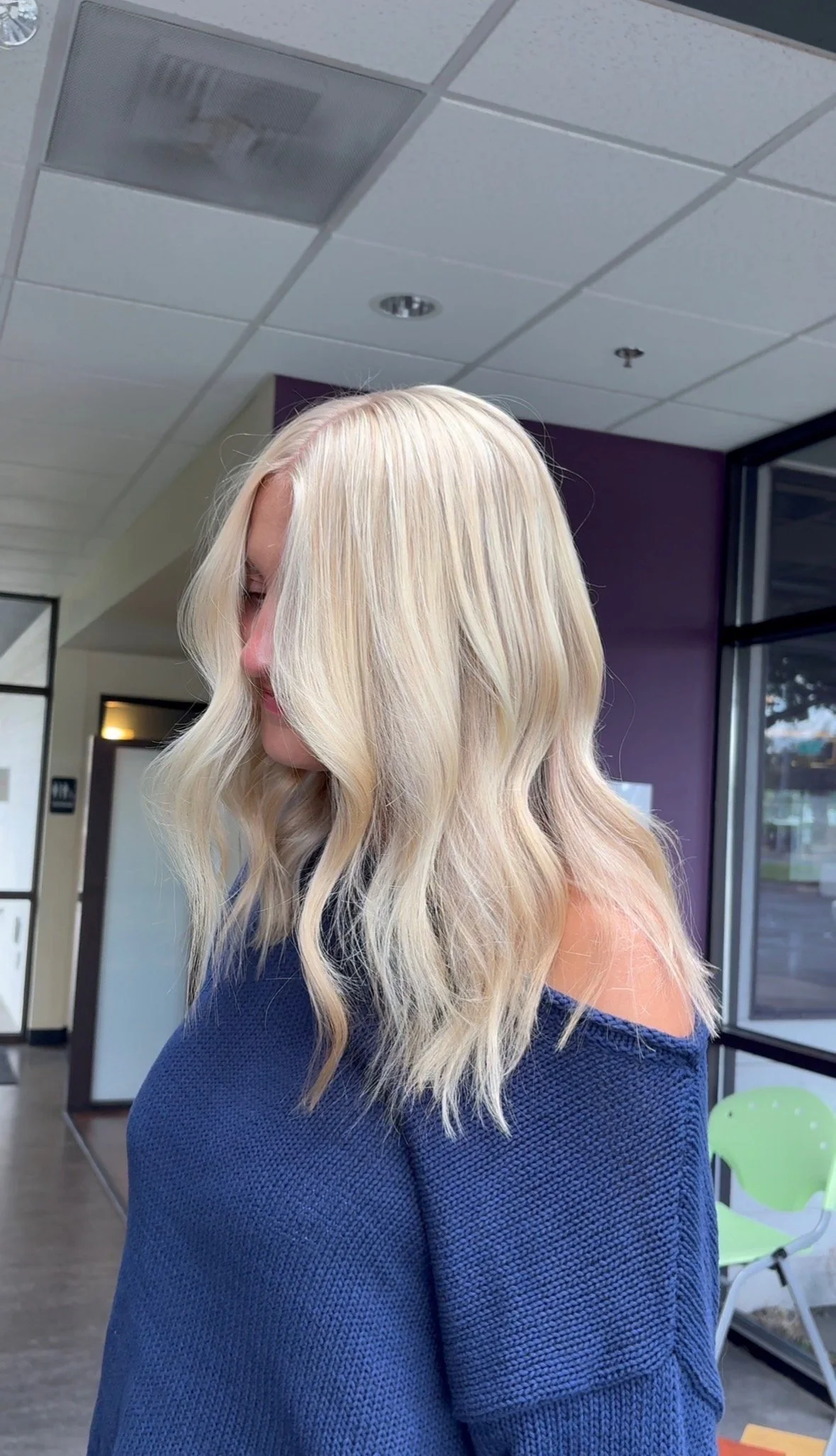 Woman with long blonde wavy hair wearing a blue cold-shoulder top, inside a modern building with large windows and a purple wall.