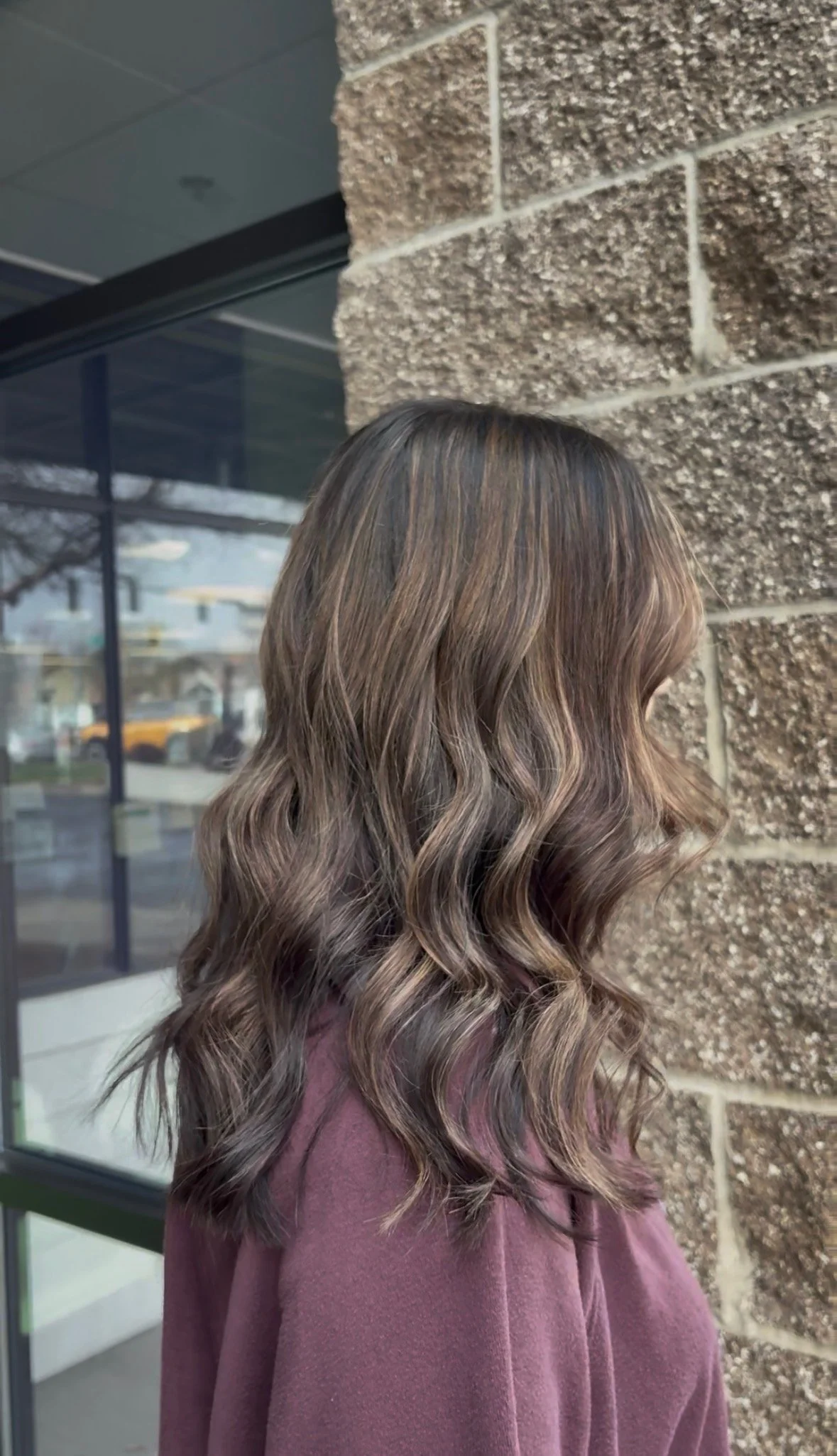 Side view of a woman with wavy, blonde hair, wearing a mauve-colored top, standing outside near a brick wall and glass window.