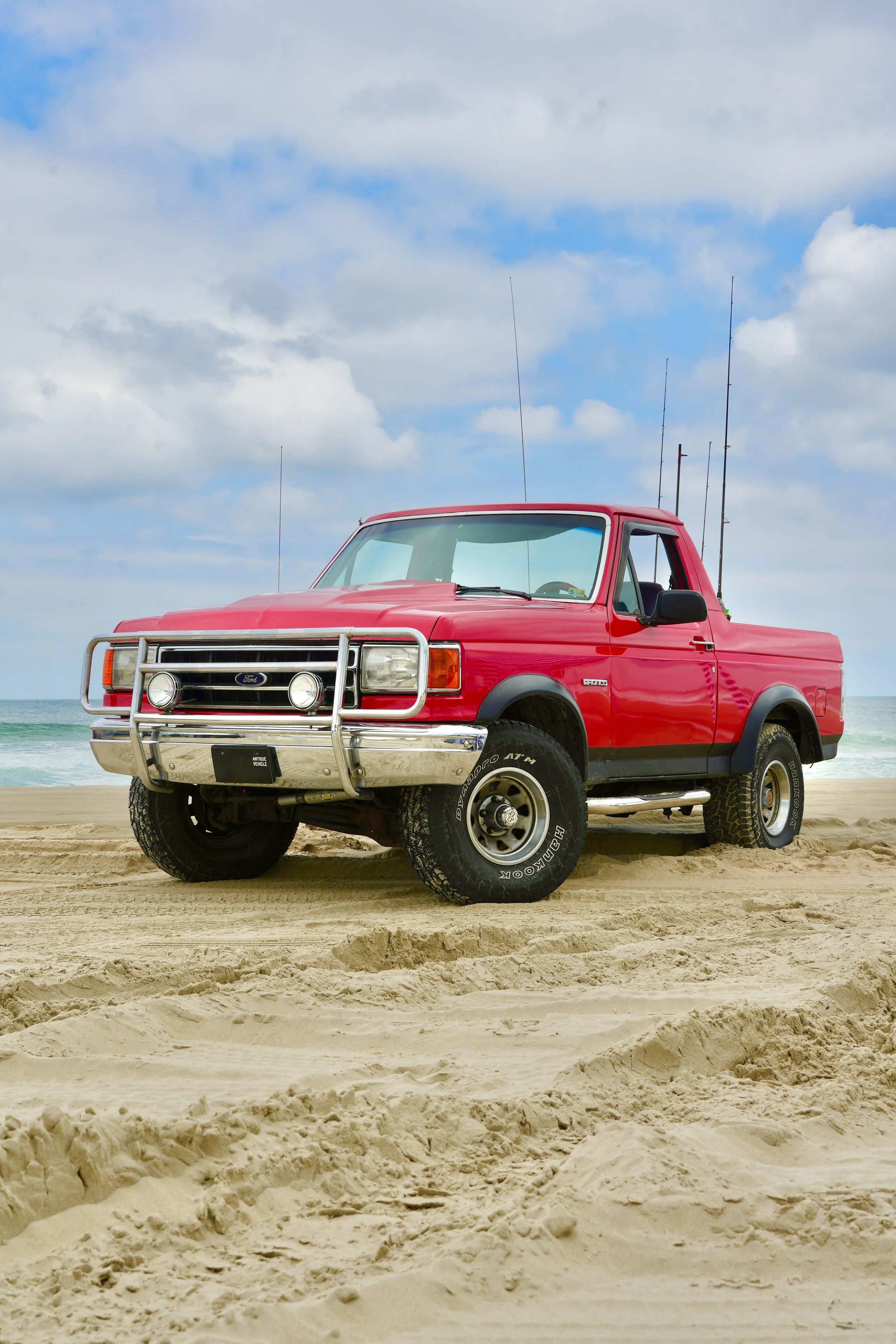 Red pickup truck parked on sandy beach with ocean and sky in the background.