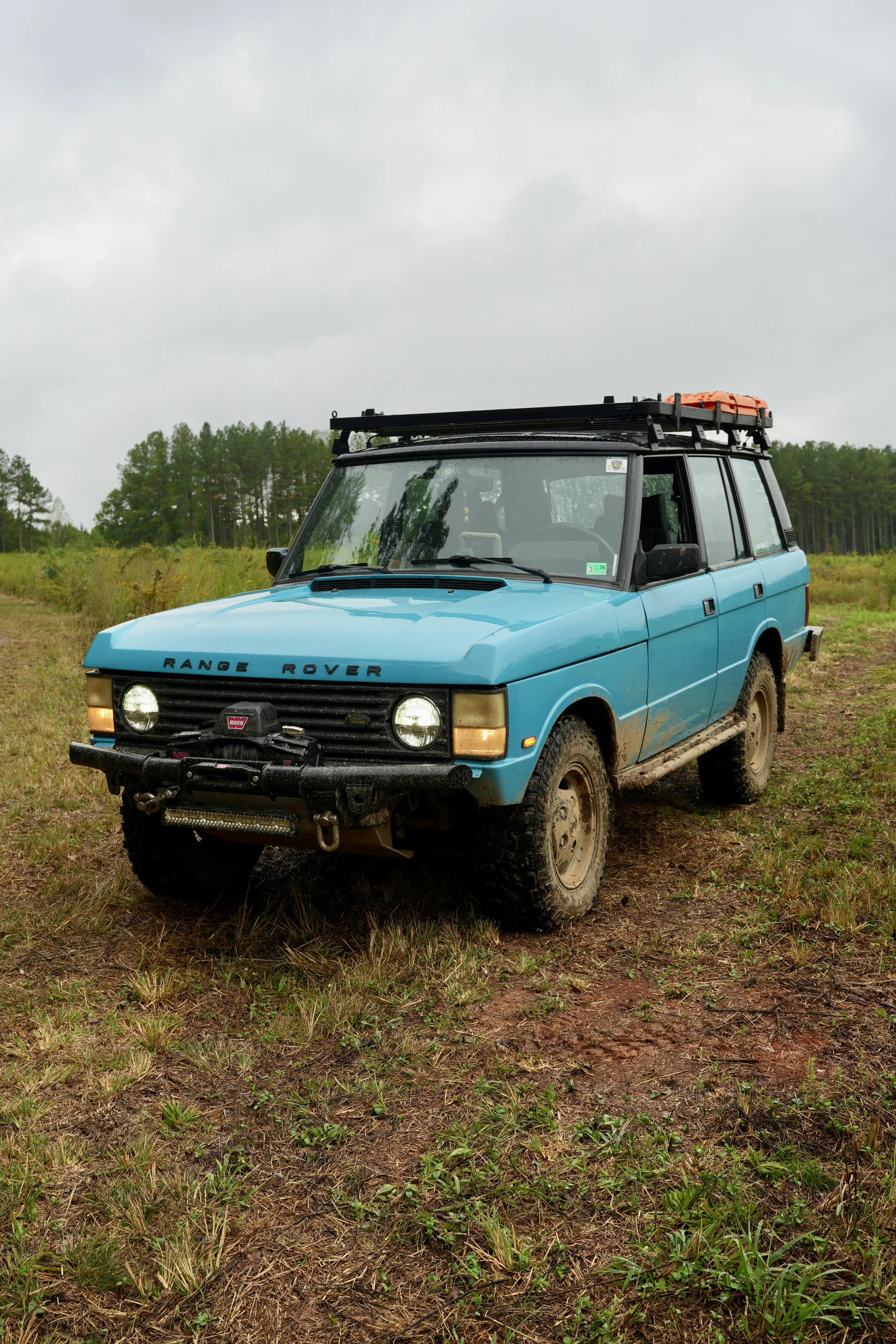 Blue Range Rover SUV parked on grassy terrain with mud on the tires, under a cloudy sky, with trees in the background.