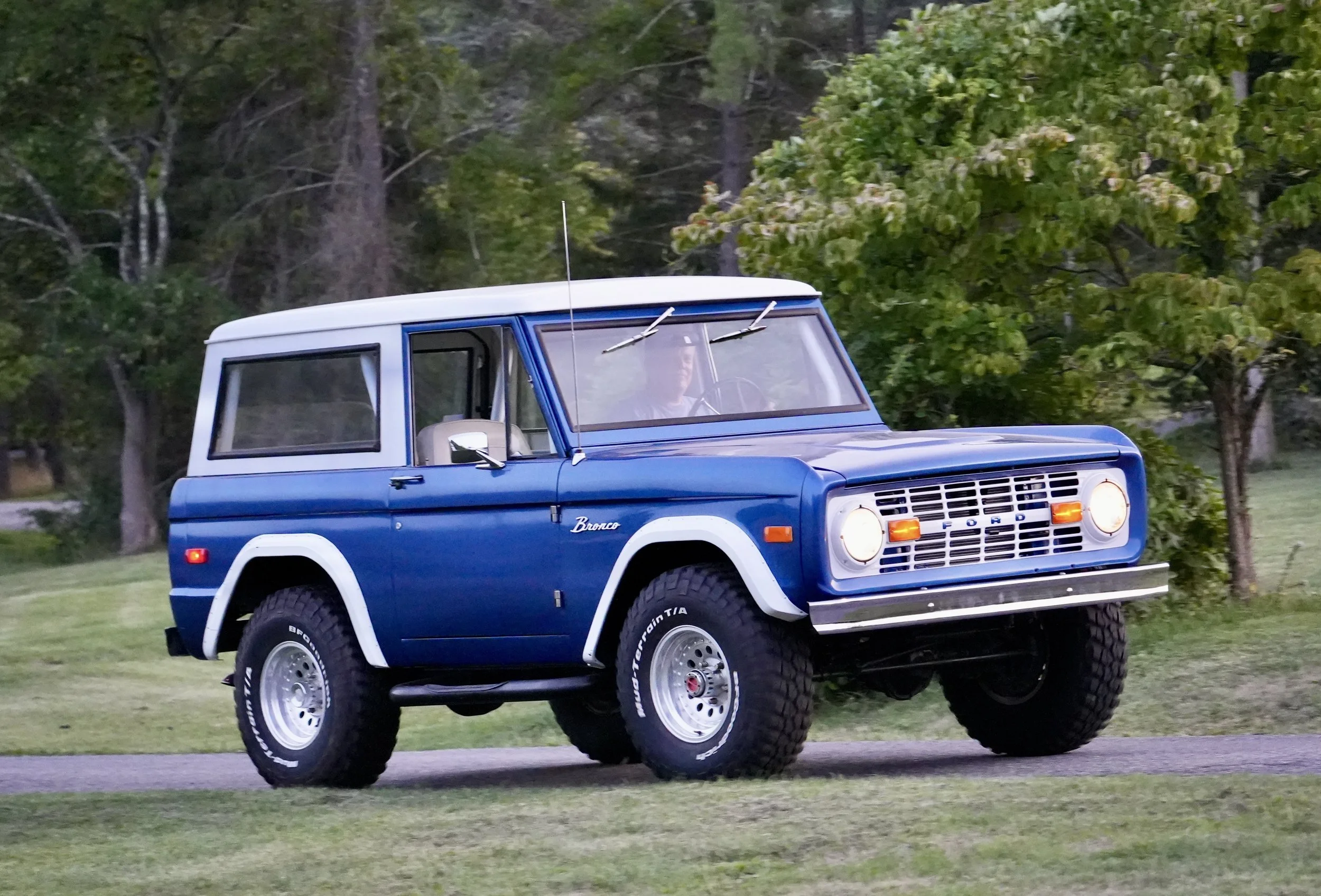A blue vintage Ford Bronco SUV driving on a grassy area surrounded by trees.