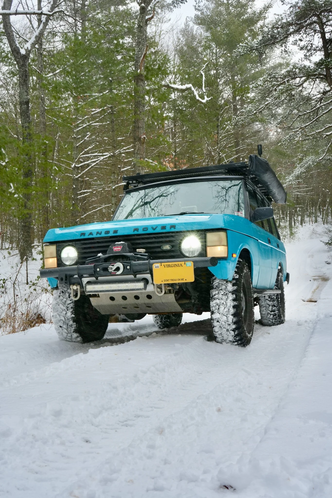 A blue Range Rover parked on a snowy ground in a rural or forested area, with snow-covered trees in the background.