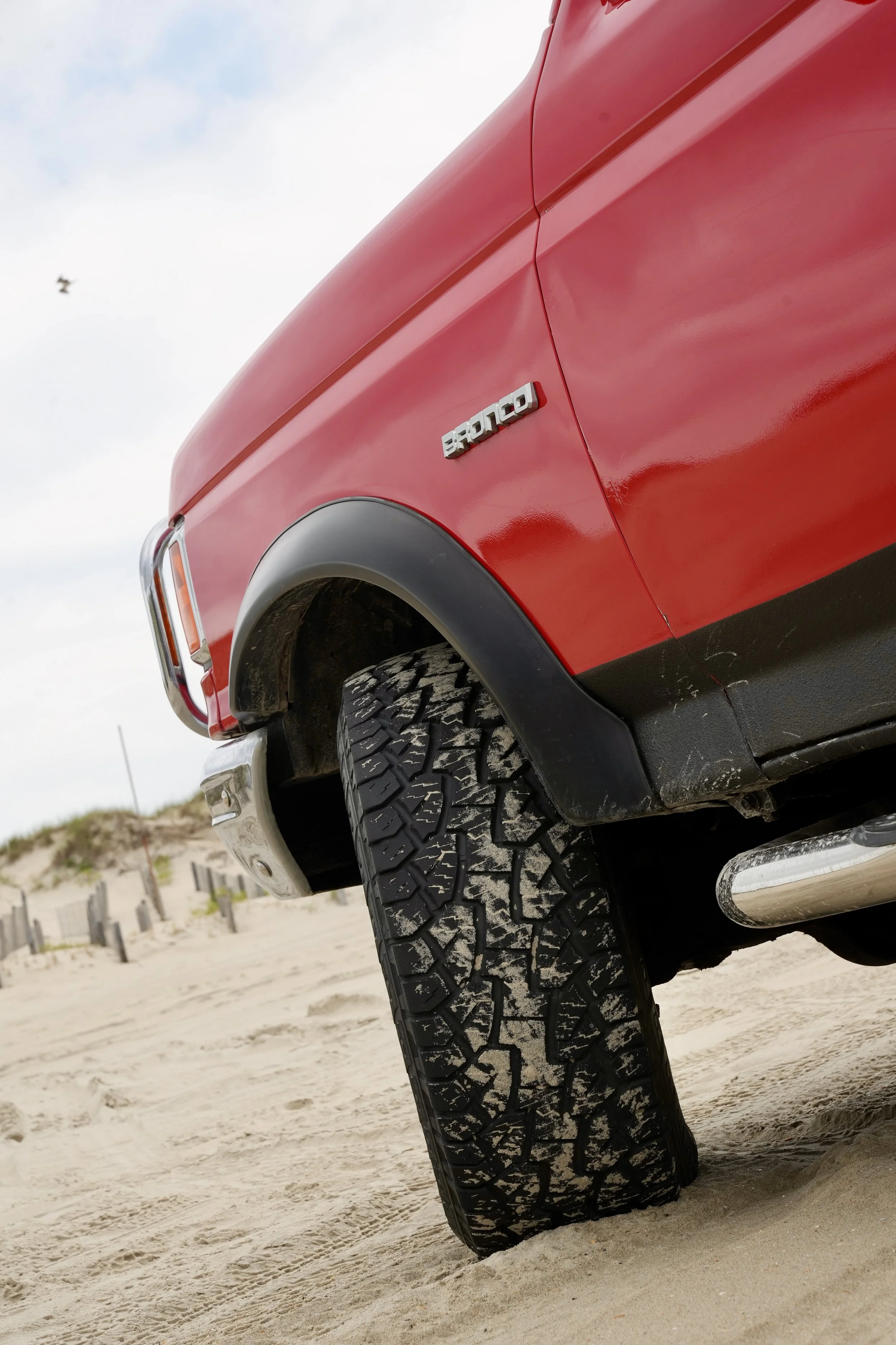 Close-up of the front side of a red pickup truck with a tire on sandy ground. The truck badge reads 'SPORT.' The background includes a cloudy sky and a fence.