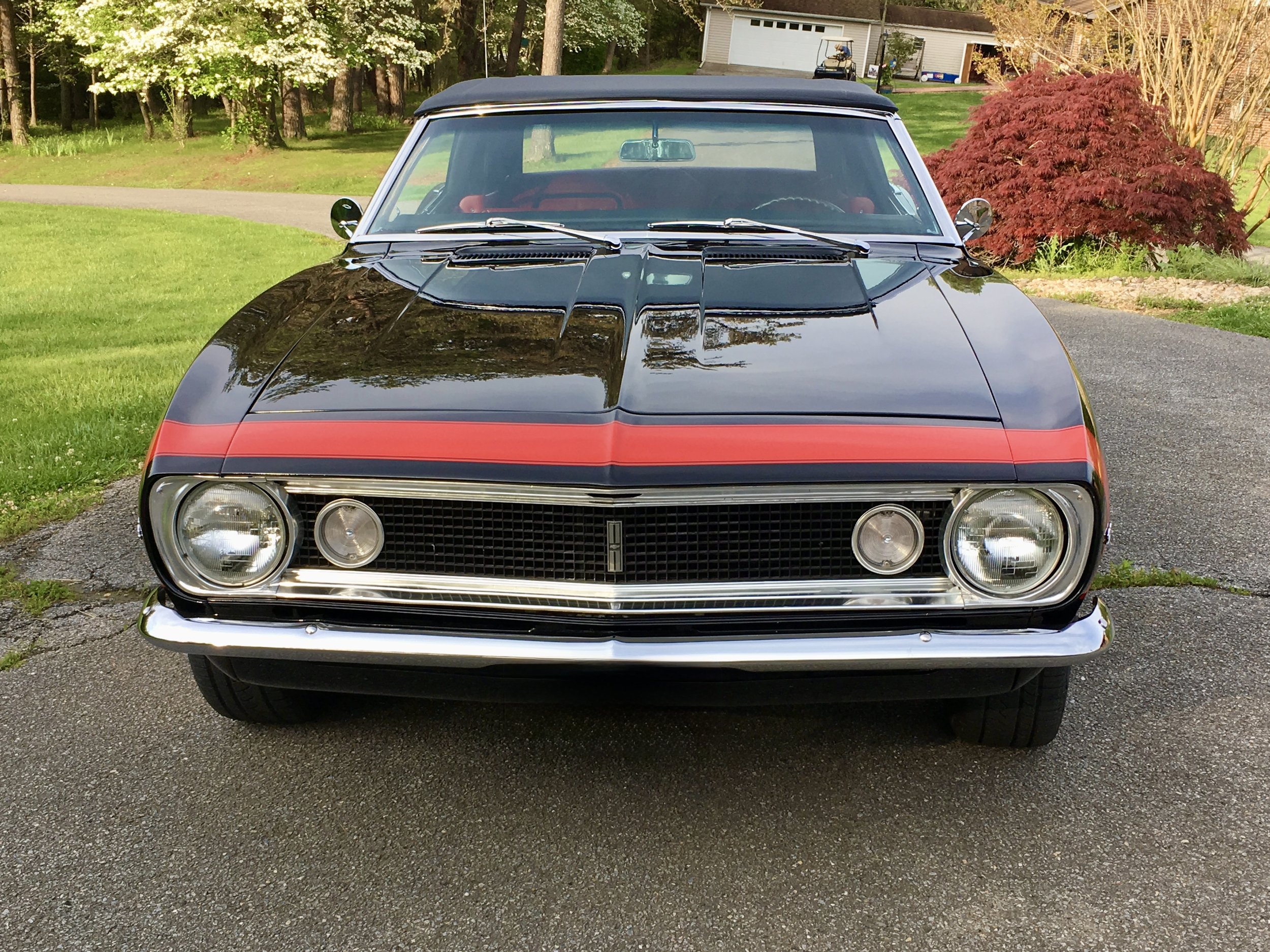 Front view of a classic black and red muscle car parked on a driveway surrounded by green grass and trees.