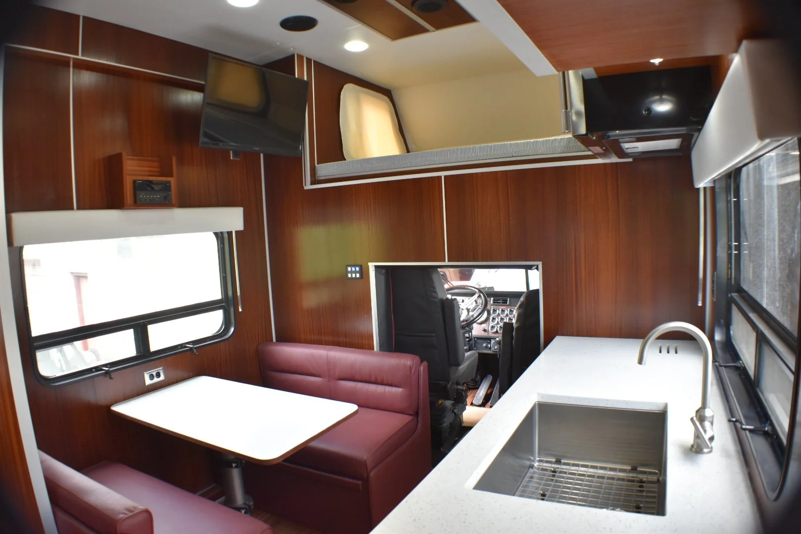 Interior of a recreational vehicle with a small dining area with a pink cushioned bench and a white table, a compact kitchen with a sink and countertop, and the driver's cab visible in the background