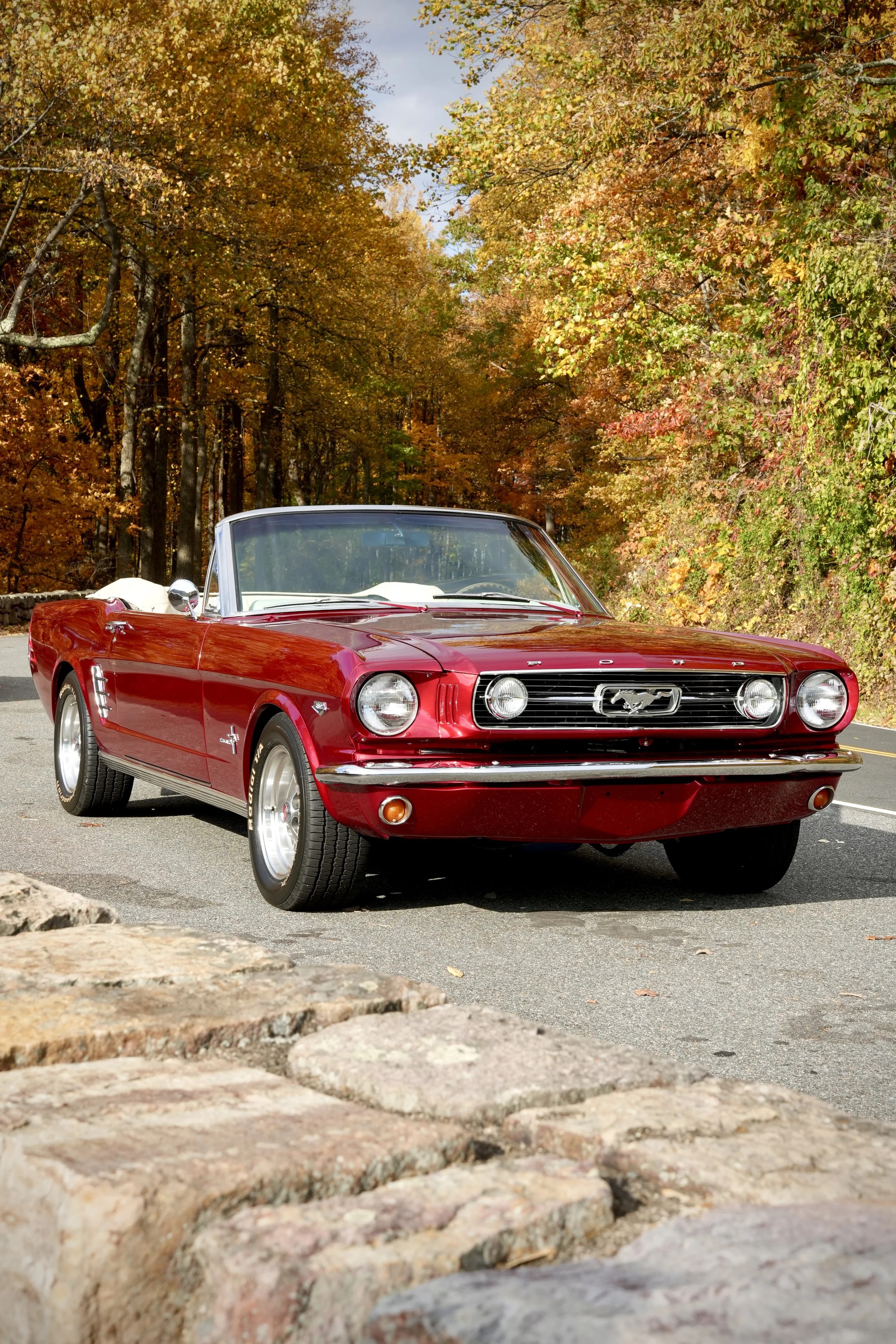 A red vintage Ford Mustang convertible car parked on the side of a road with autumn foliage in the background.