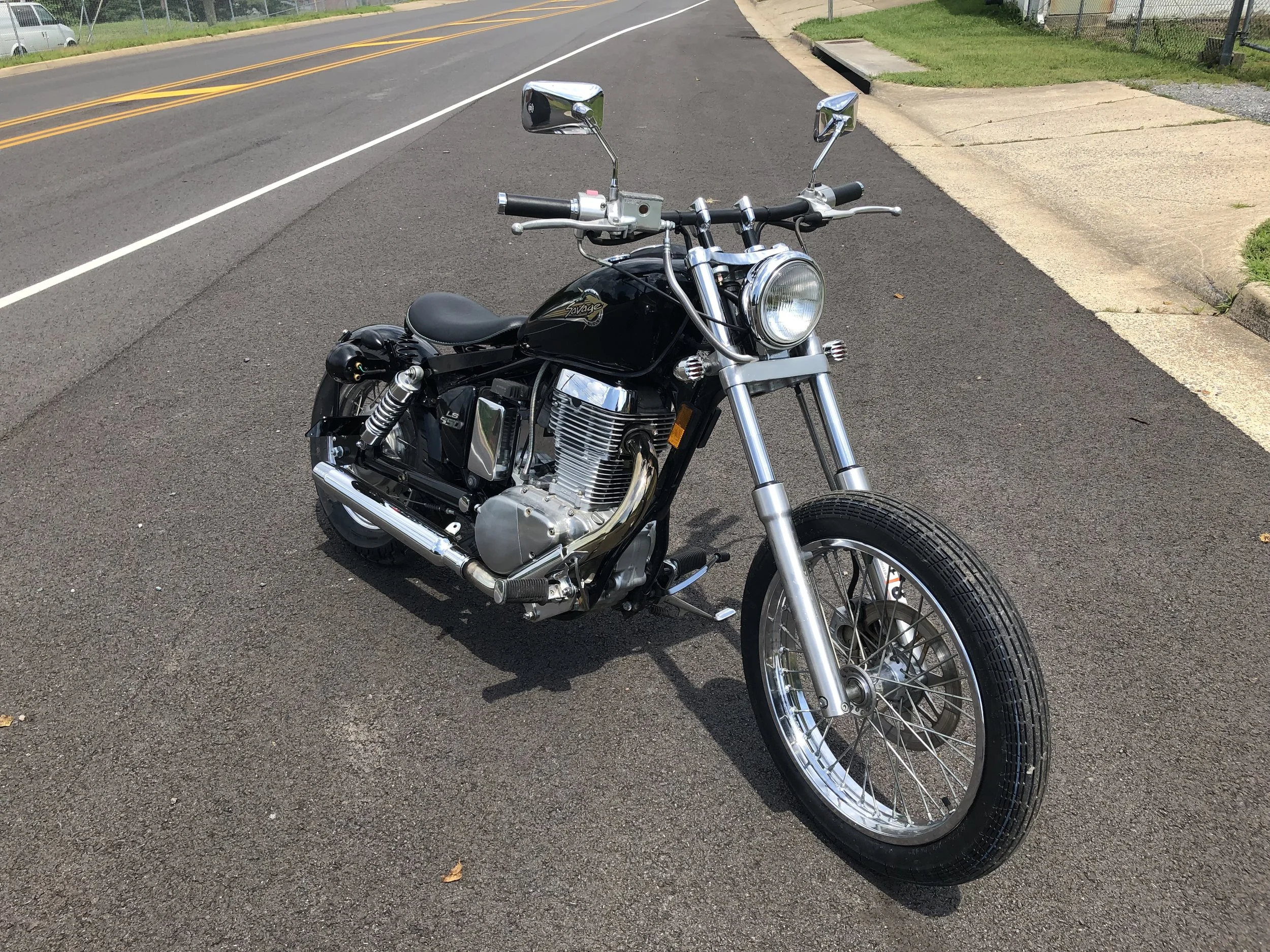 Black motorcycle parked on an asphalt road near a sidewalk, with a grassy area and a chain-link fence in the background.