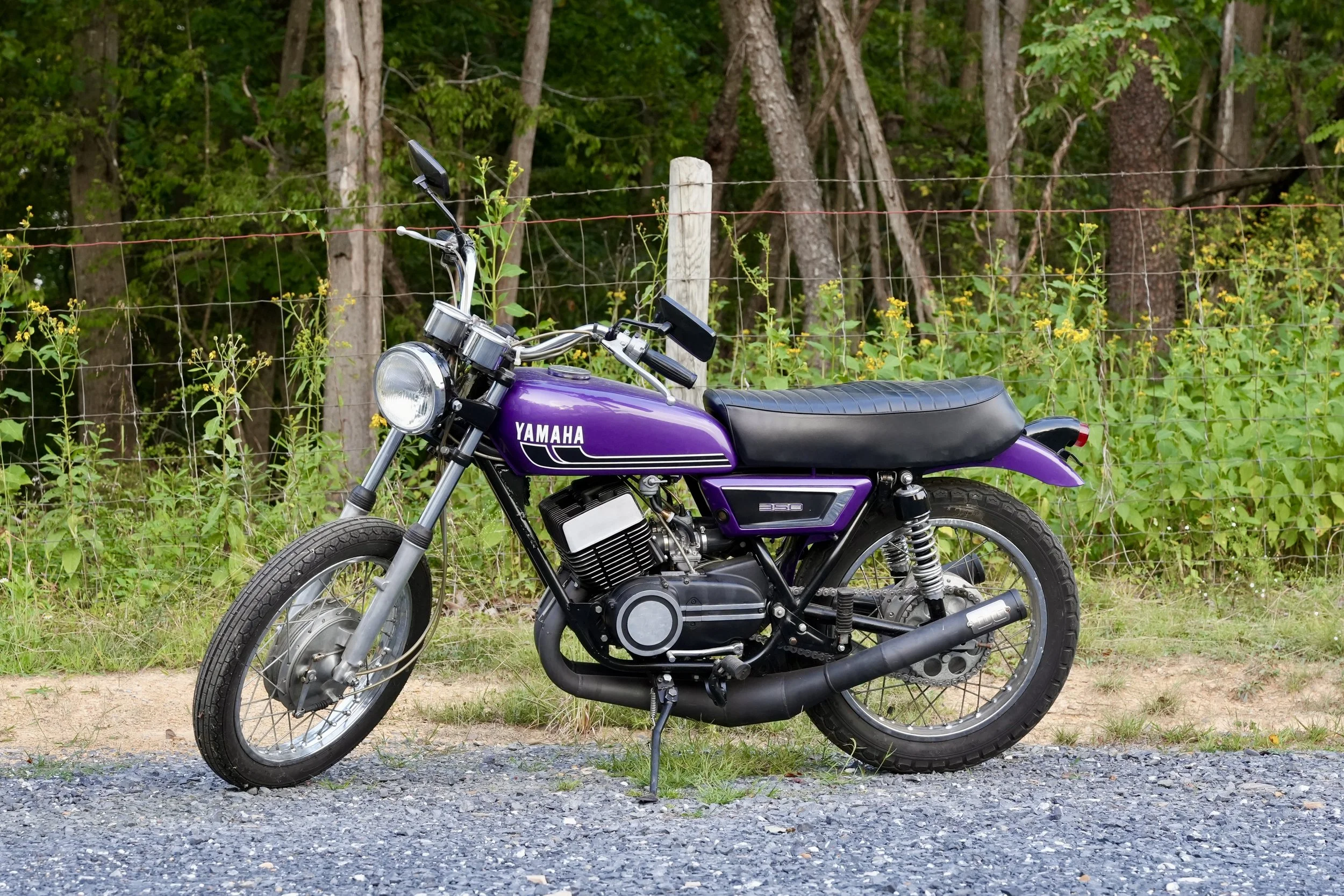 A purple Yamaha motorcycle parked on a gravel area beside a dirt path, with a wooden fence and green trees in the background.