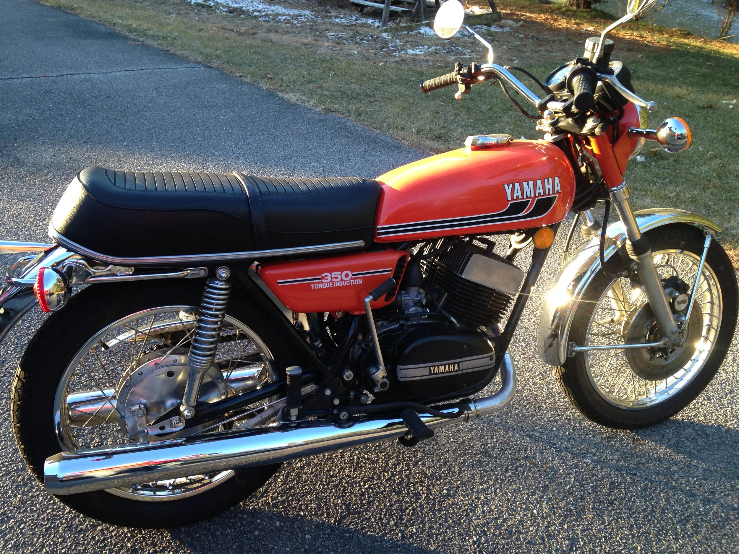 A vintage red Yamaha motorcycle parked on a paved road with grass and trees in the background, illuminated by sunlight.