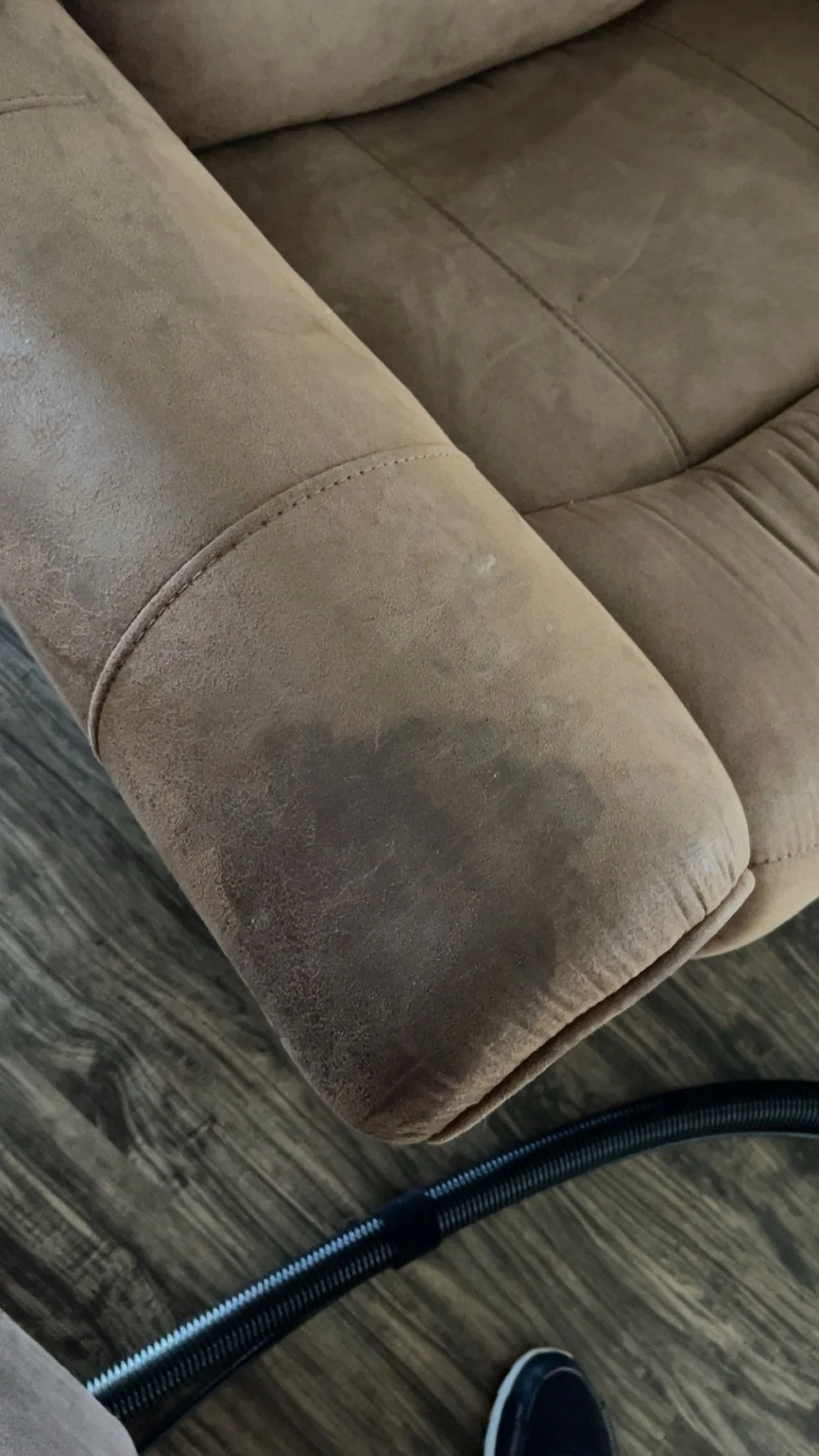 Close-up of a beige fabric sofa and a person’s sneaker on a hardwood floor, with a black vacuum hose and a coiled black cable on the floor.
