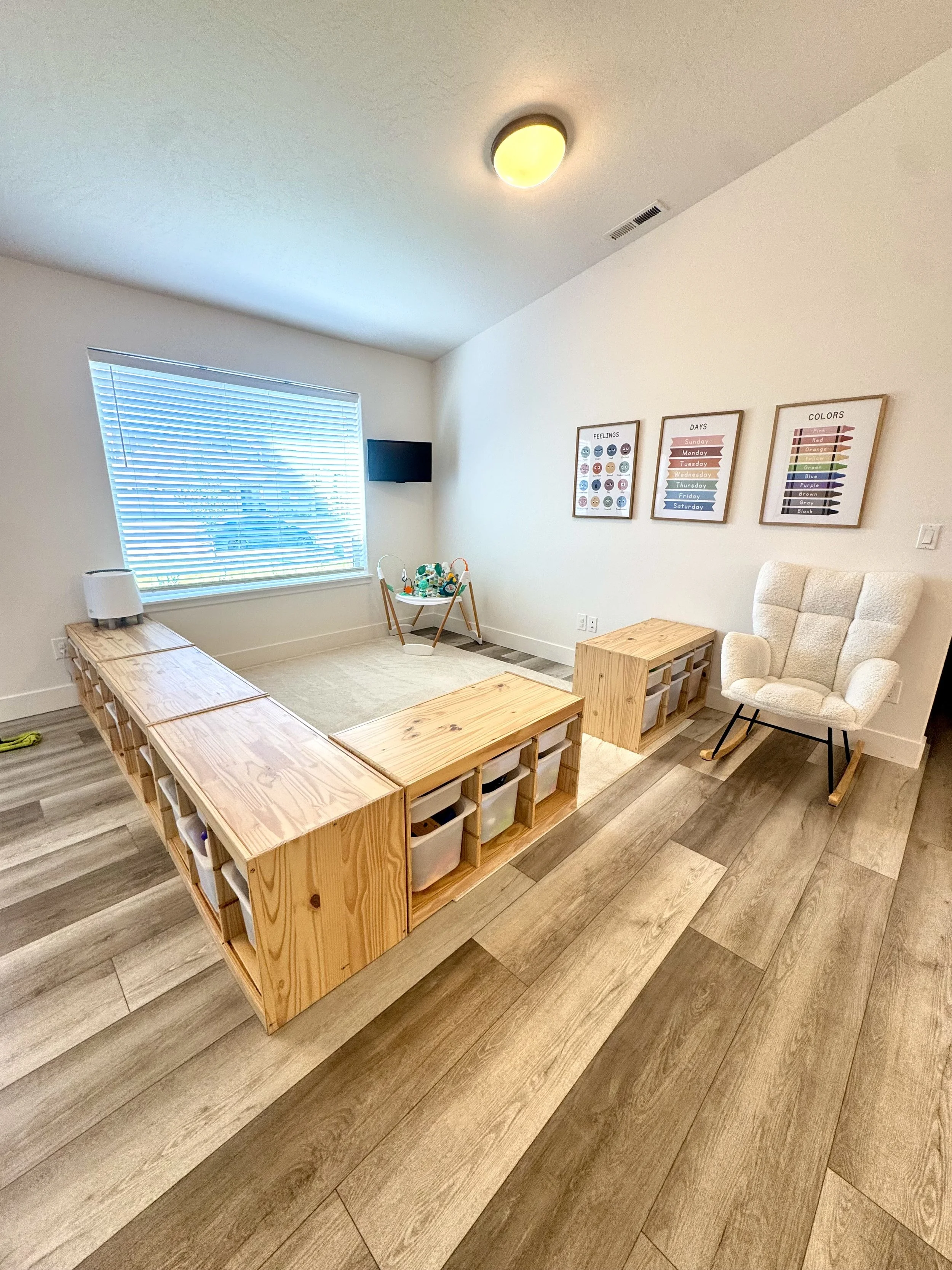 A cozy living room with a large window with blinds, a beige armchair, two wooden coffee tables, a white side table, and framed artwork on the wall featuring feelings, days, and colors. The room has wood flooring and minimal decor.