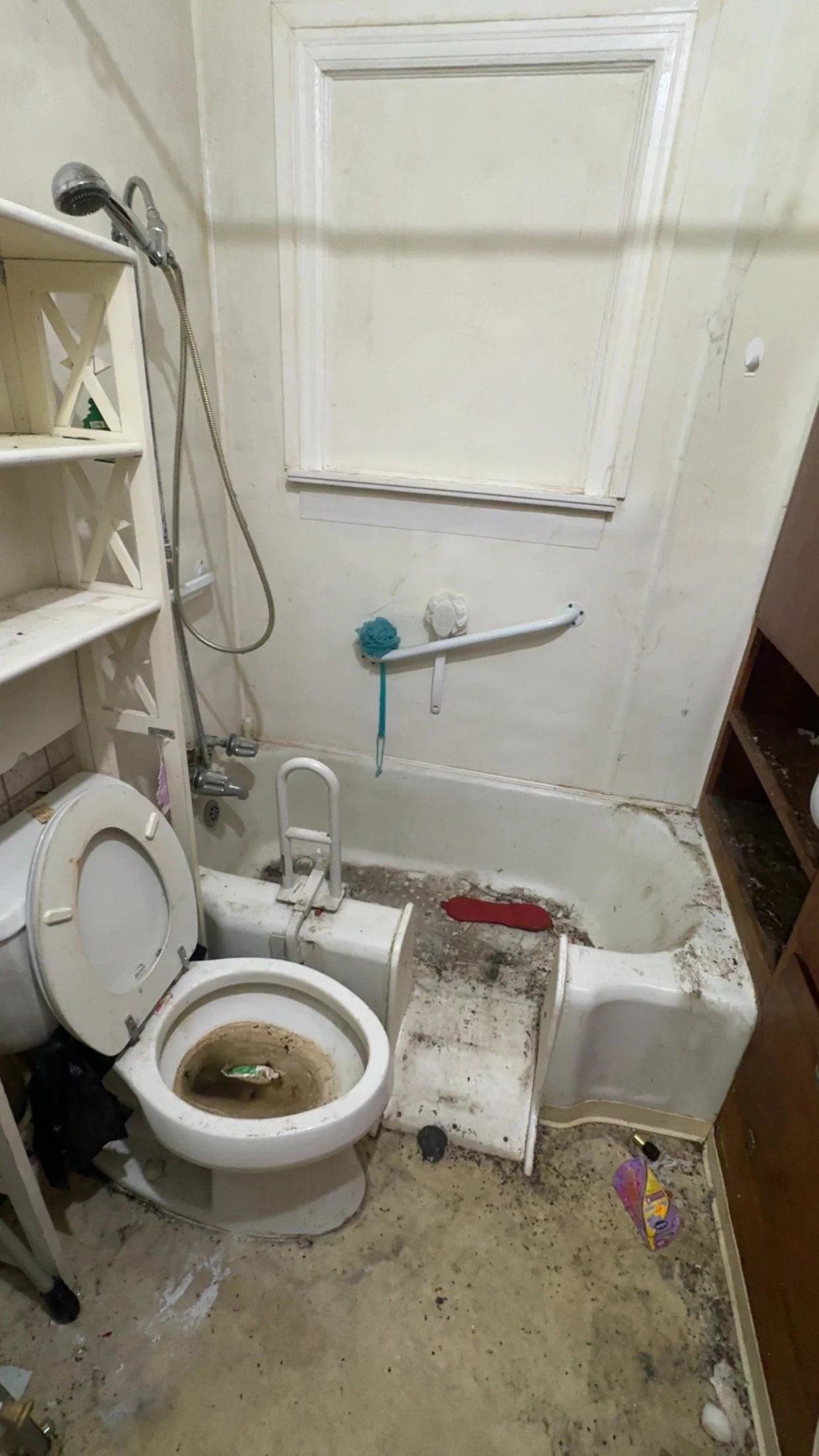 A dirty, broken bathroom with a stained toilet, exposed plumbing, peeling paint, and scattered debris.