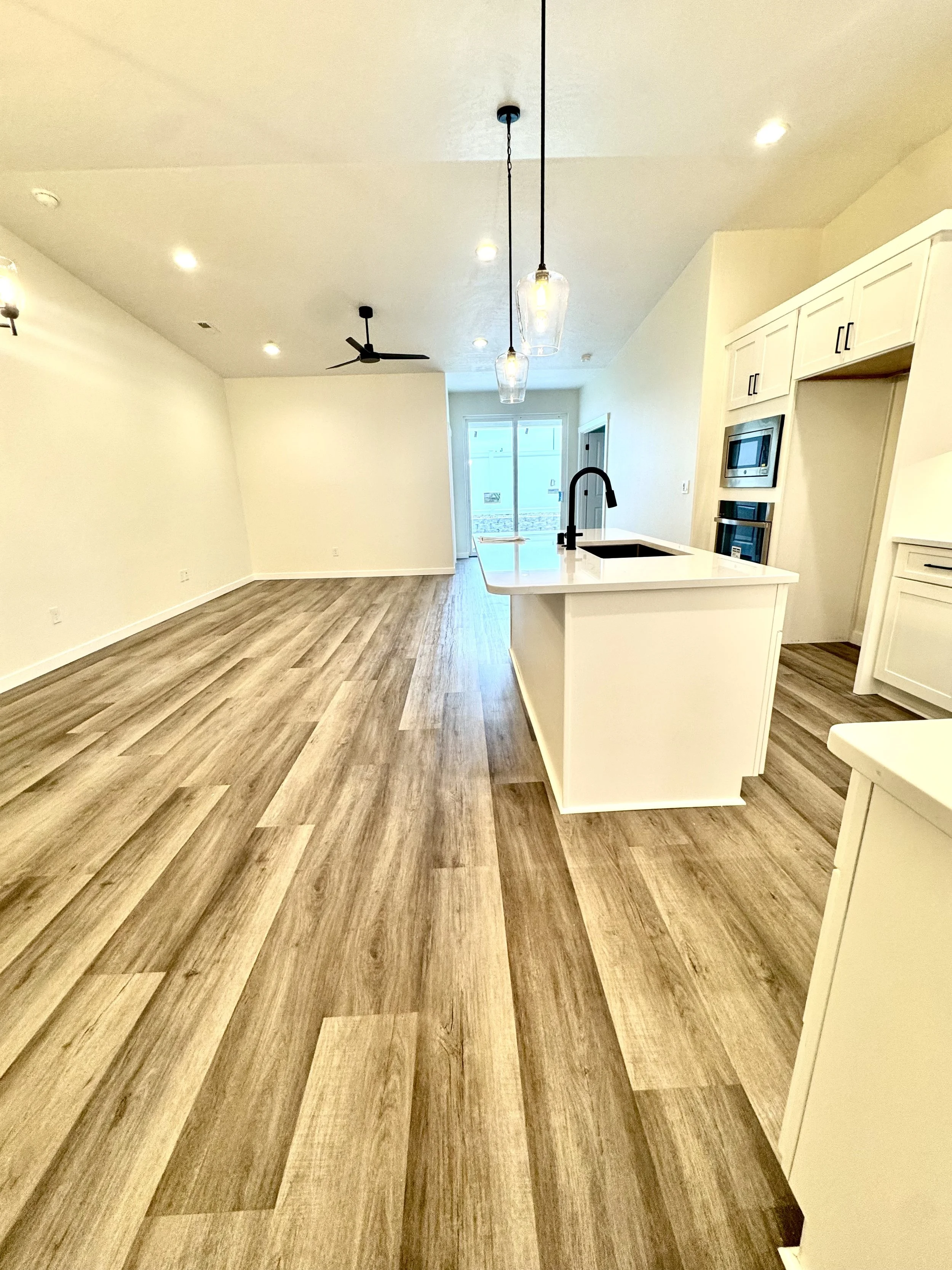 Open-concept kitchen and living area with wood flooring, white cabinetry, a kitchen island with a black sink and faucet, pendant lights, and a sliding glass door leading outside.