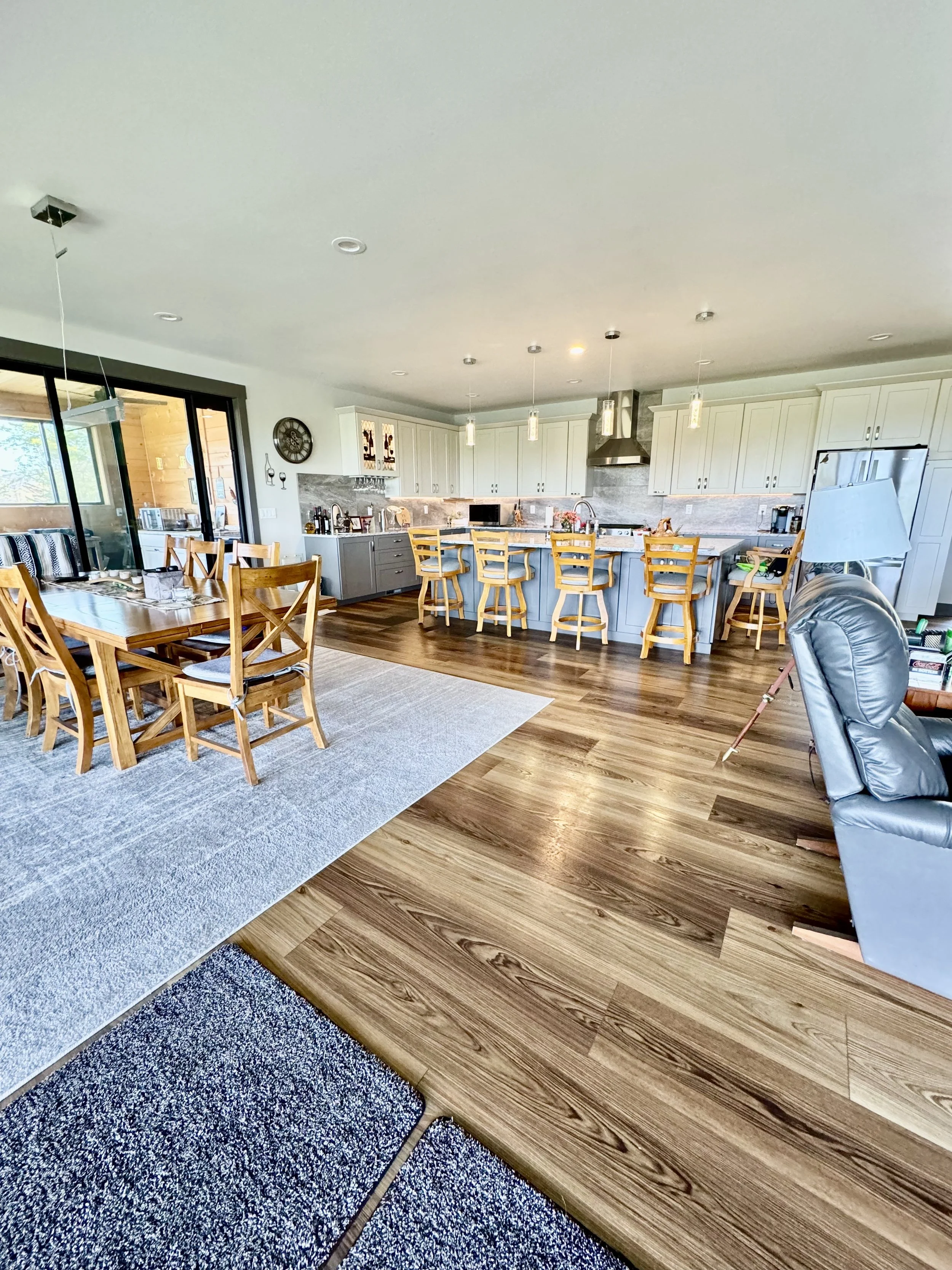 Photo of a modern kitchen and dining area with wooden flooring, white cabinets, a kitchen island with barstools, and a dining table with chairs.