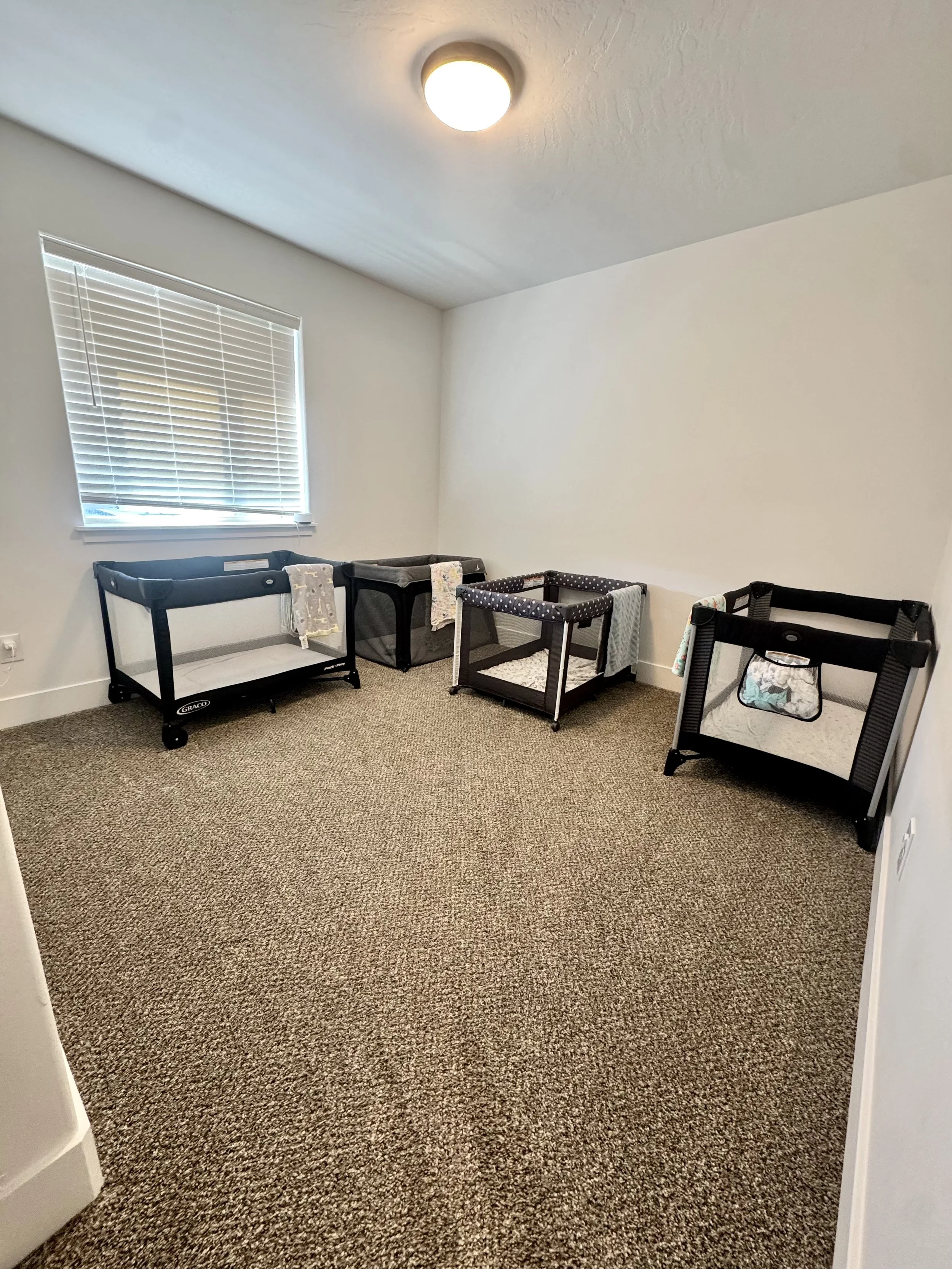 A room with beige carpet flooring and white walls. There are four playpens with fabric sides placed against the wall near a window with white blinds. The room is lit by a ceiling light.