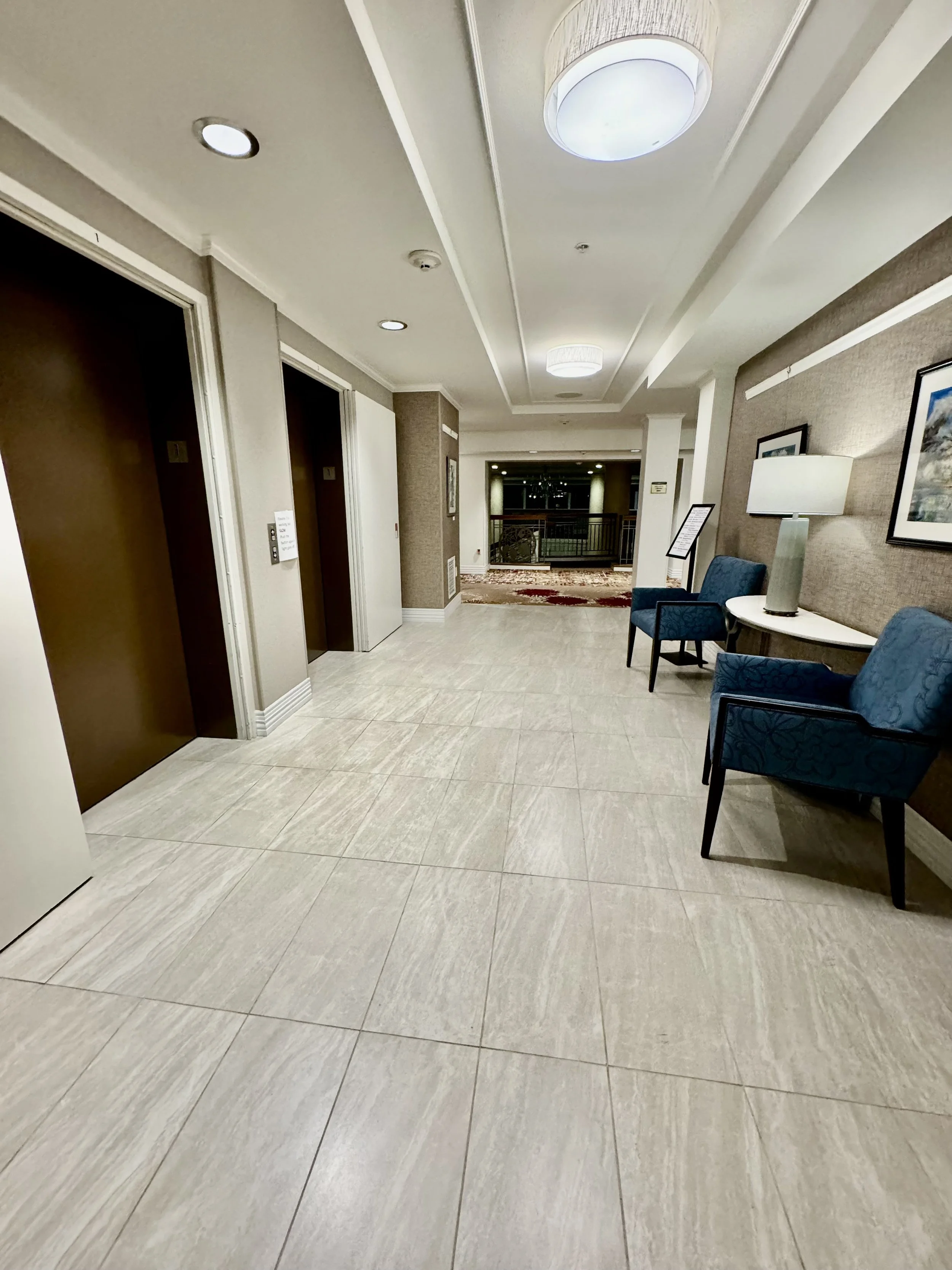 Lobby area with two elevator doors on the left side, seating with blue chairs and a side table with a lamp on the right, framed pictures on the wall, and a hallway with a railing at the end.