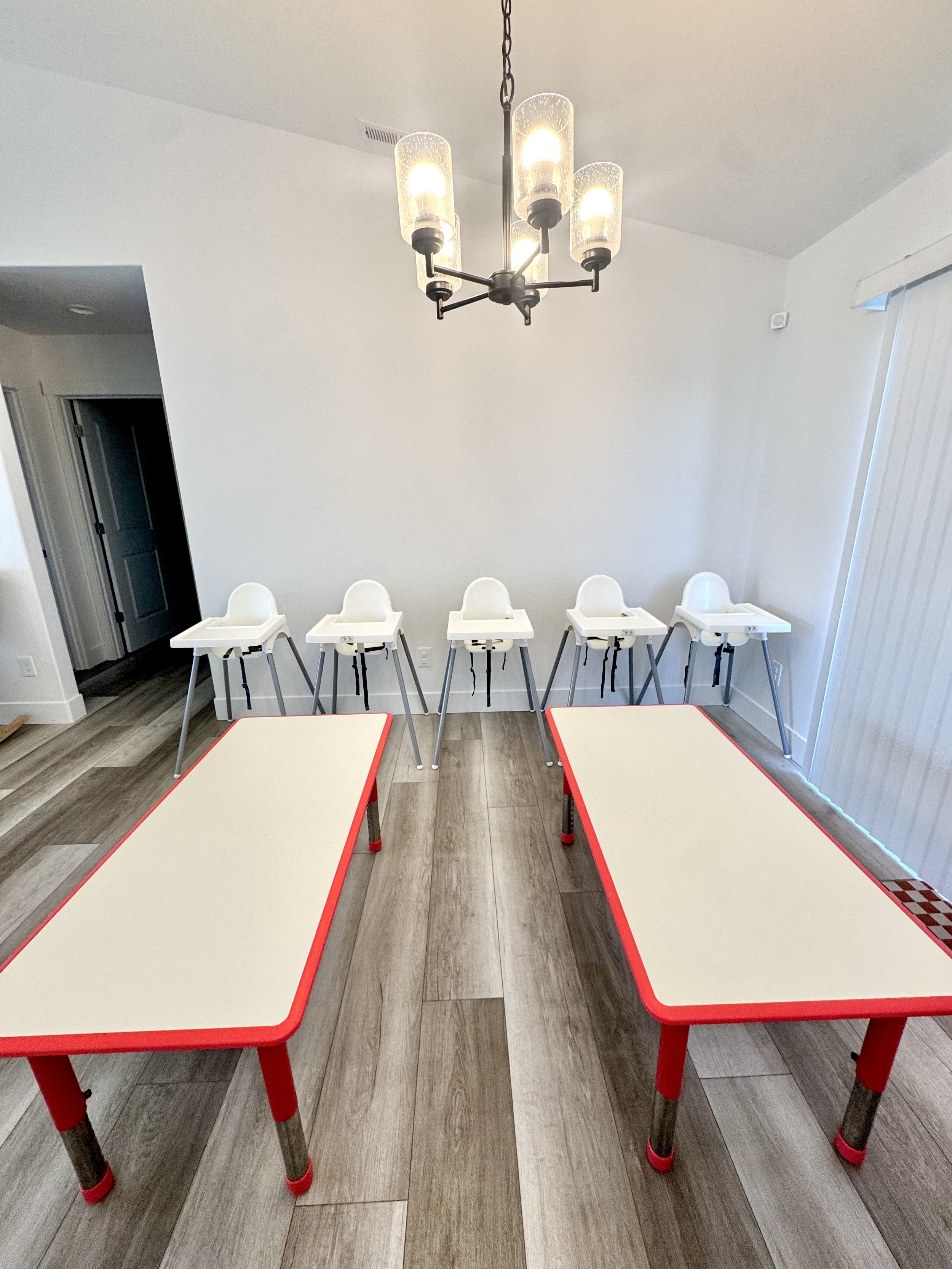 Five white high chairs line up against a white wall, with two children's tables in the foreground, and a modern chandelier hanging from the ceiling as part of a childcare or dining area.