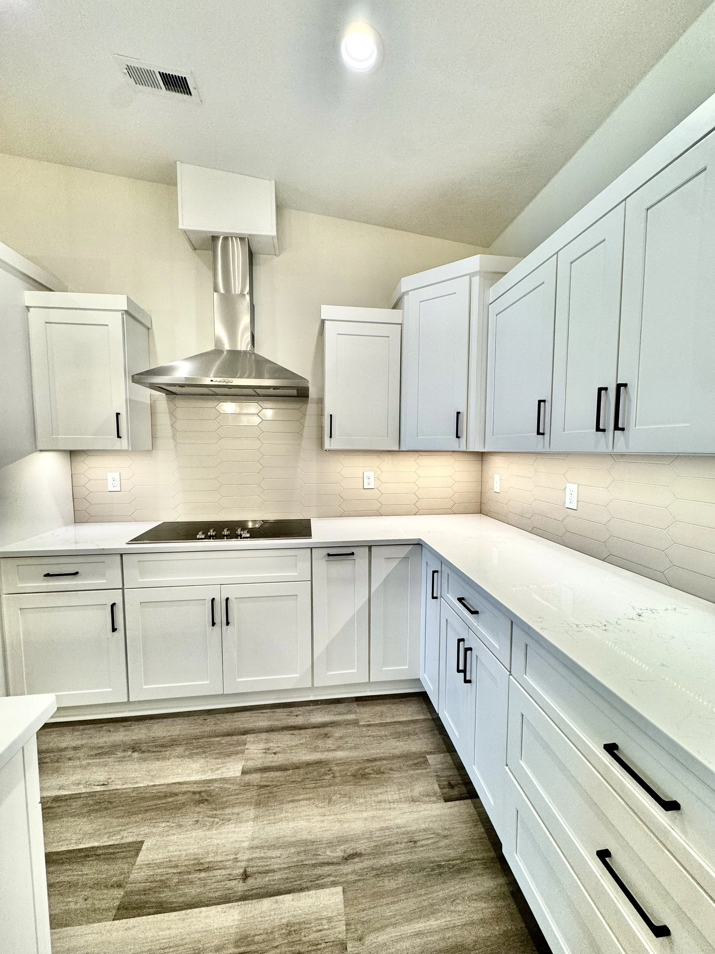 Modern kitchen with white cabinets, a stainless steel range hood, hexagonal tile backsplash, dark hardware, and wood-style flooring.