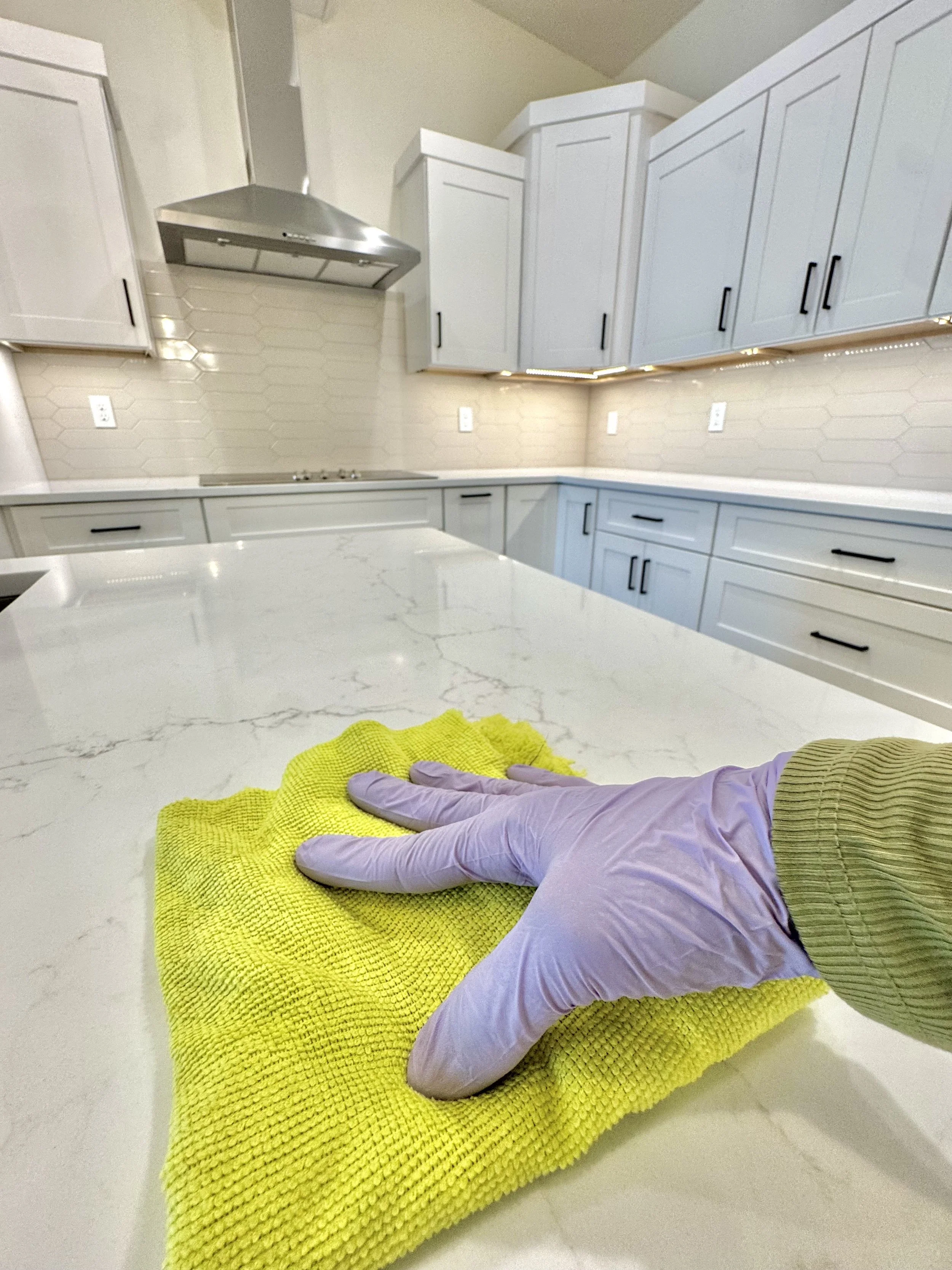 View of a person's arm and hand wearing a purple sleeve and a yellow glove, resting on a yellow textured cloth on a white kitchen countertop. The background shows white cabinets, a kitchen range hood, and hexagonal tile backsplash.