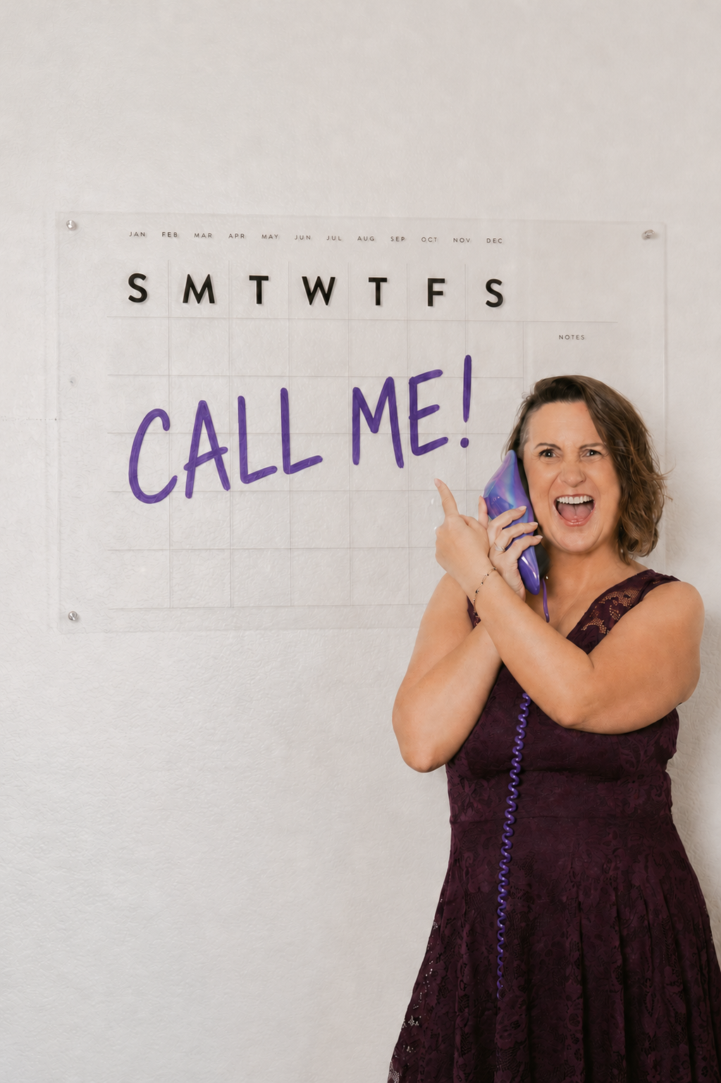 Kim Ast A wearing a purple dress is talking on a corded landline phone with a surprised or excited expression, in front of a calendar on a white wall with words "Call Me!" written in large purple letters.