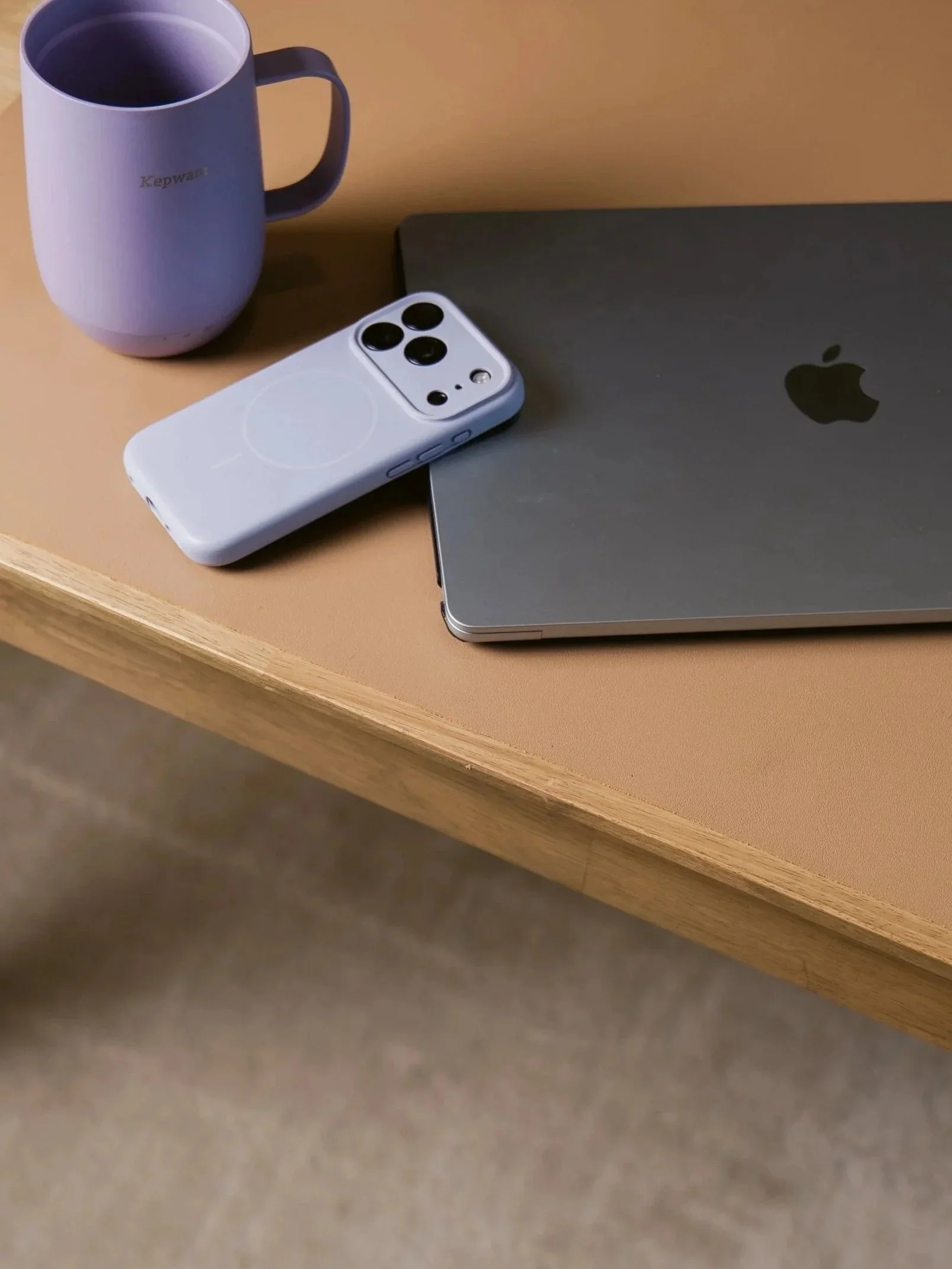Close-up of a gray laptop, a white smartphone with a protective case, a purple mug with hot beverage, all placed on a wooden desk.