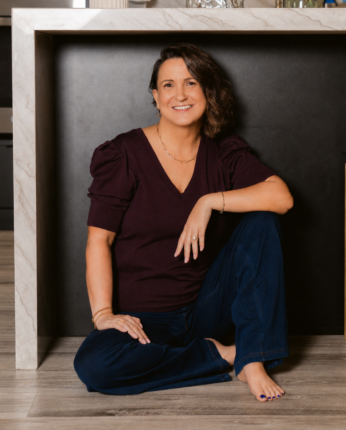 Kim Ast with shoulder-length dark hair smiling, sitting cross-legged on a wooden floor under a marble countertop.