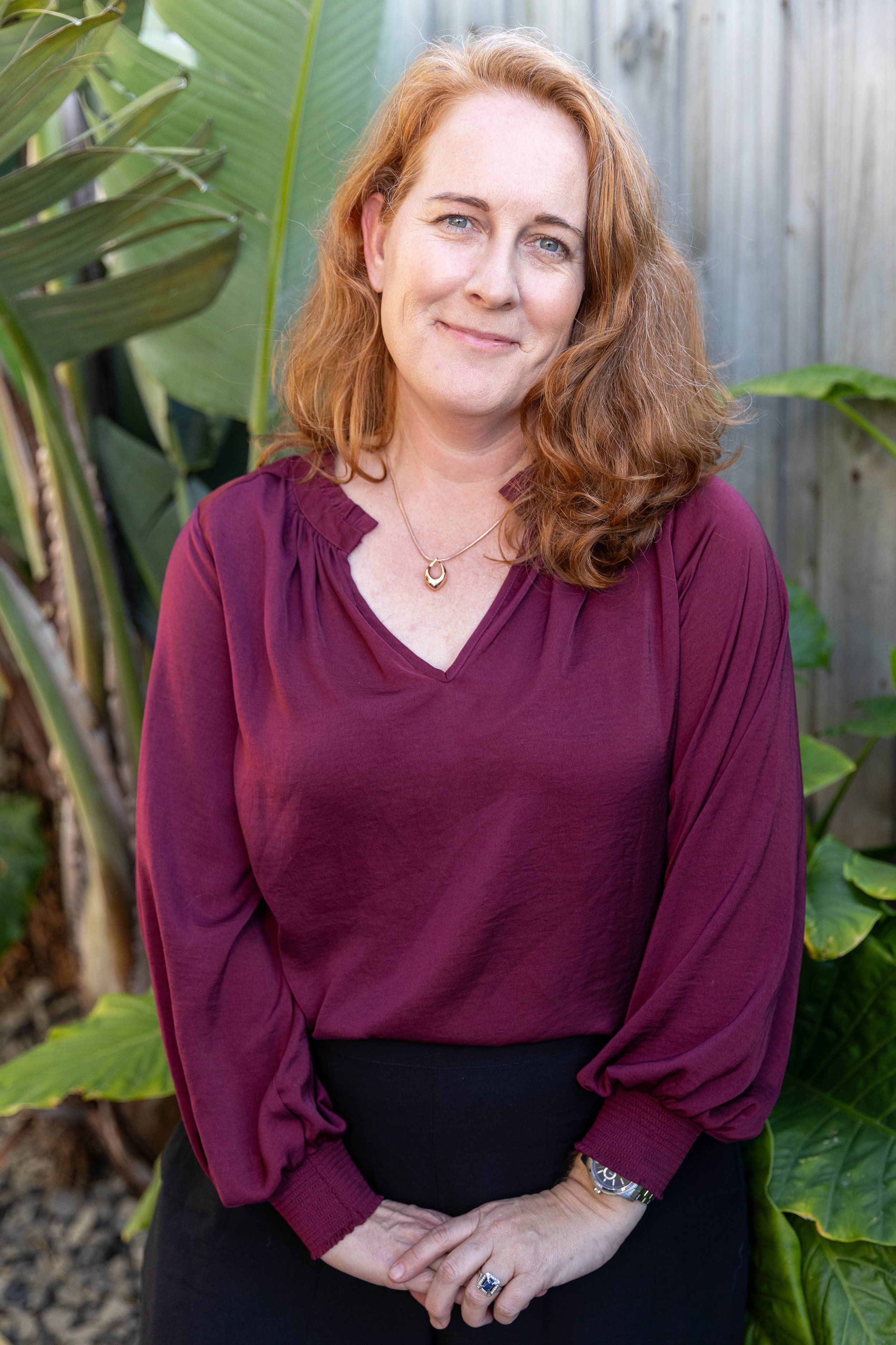 Smiling red-haired woman in a burgundy blouse and necklace posing among green plants