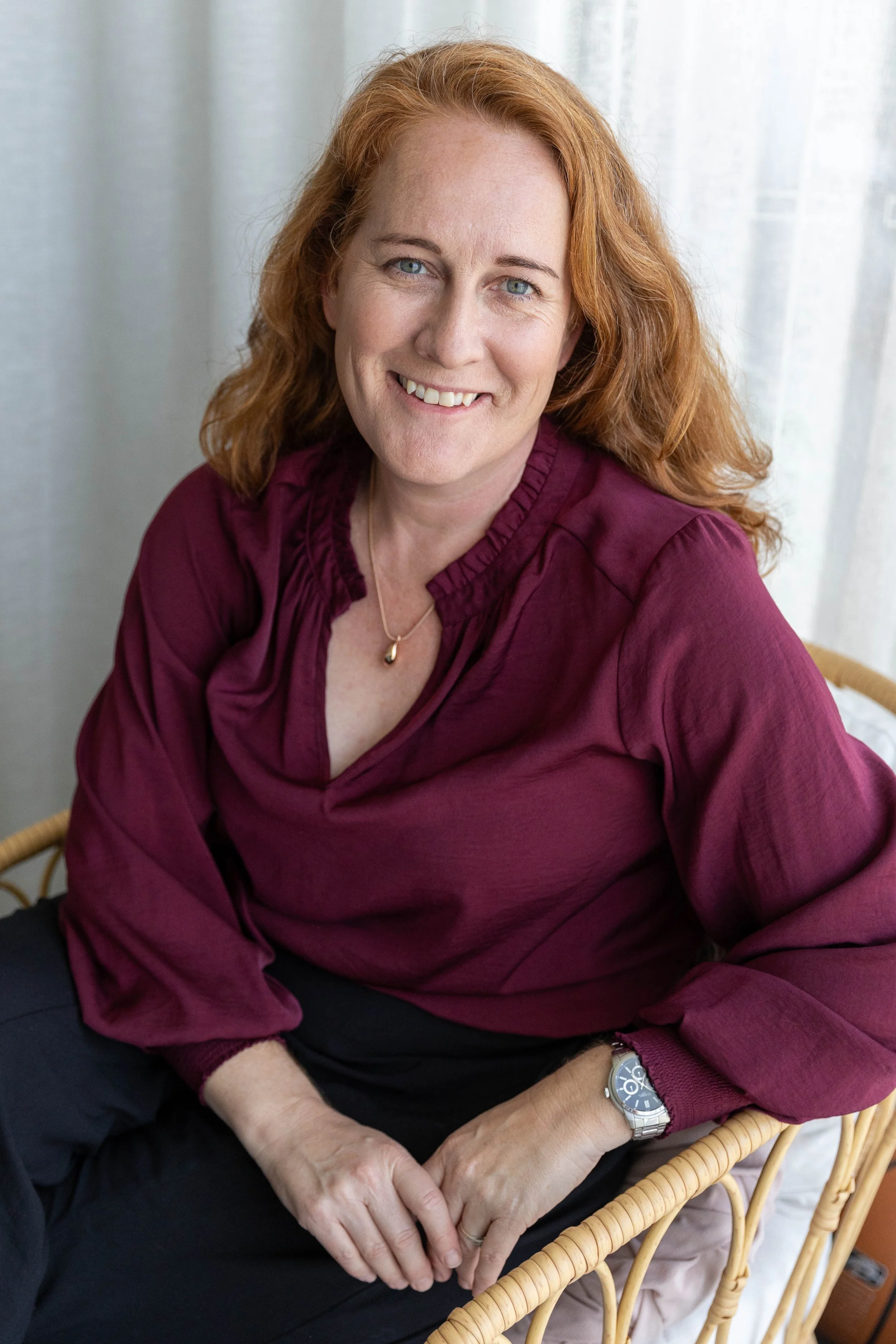 Smiling red-haired woman in a burgundy blouse seated in a wicker chair indoors