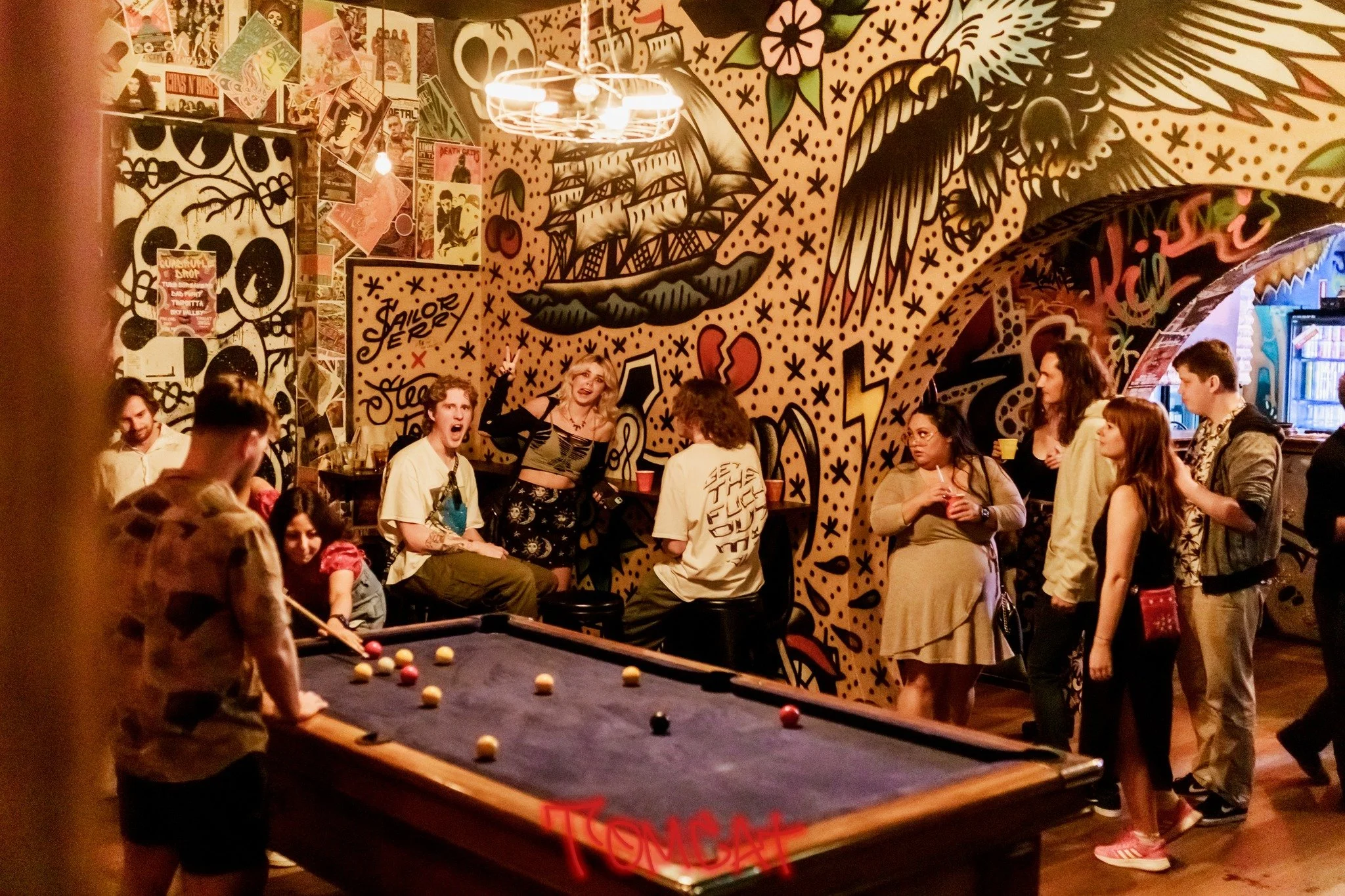 People gathered in a lively bar covered with graffiti, drinking and playing pool.