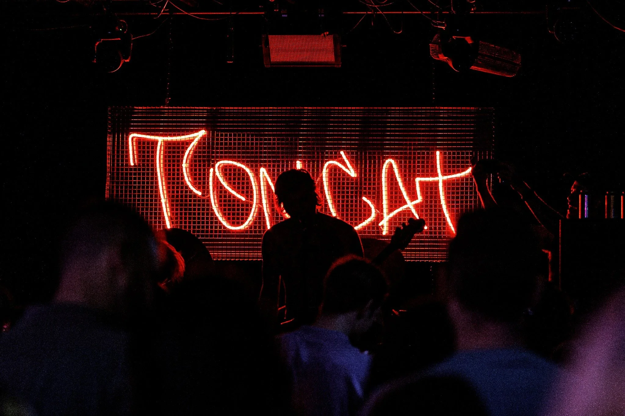 A dark scene with a red neon sign that reads 'Tomcat' and a silhouettes of a guitarist in front of it.