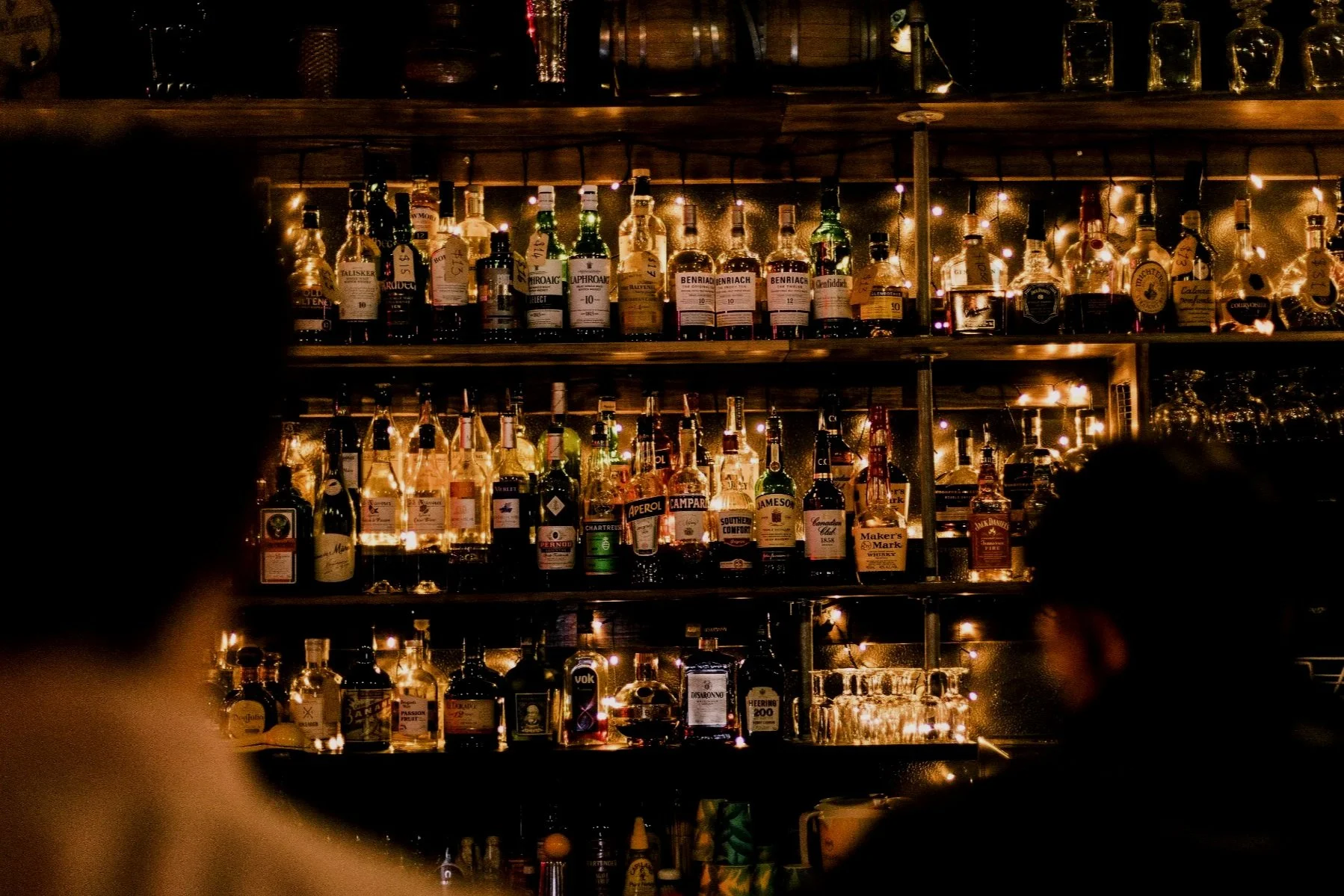 A dimly lit bar shelf with multiple bottles of alcohol, including whiskey and liquor, arranged in rows with warm string lights illuminating them.