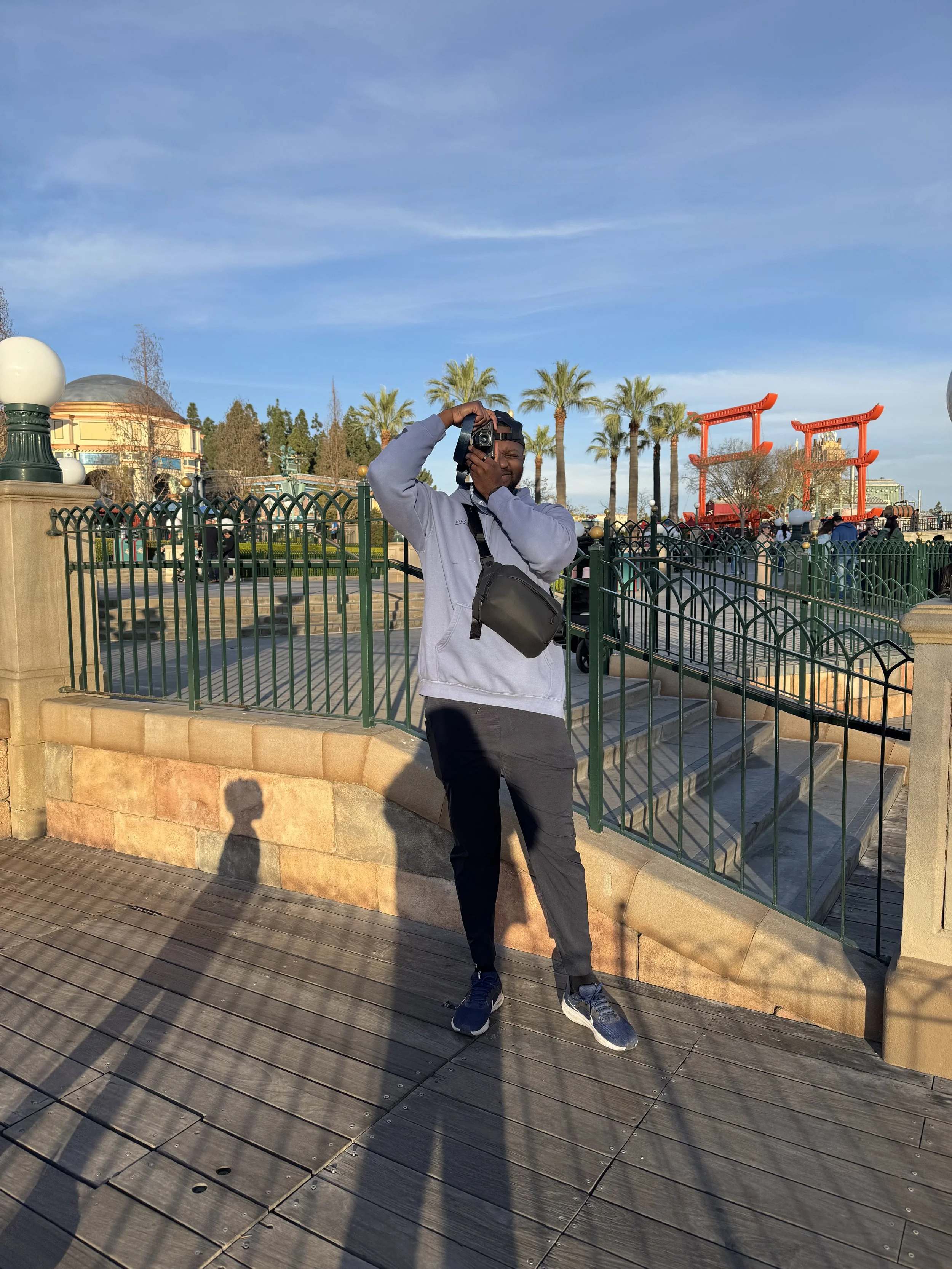 Young man in gray hoodie and black pants taking a selfie with a camera at an outdoor park area. Background includes palm trees, orange torii gates, and other visitors. The scene is lit by late afternoon sunlight, with shadows on the wooden deck.
