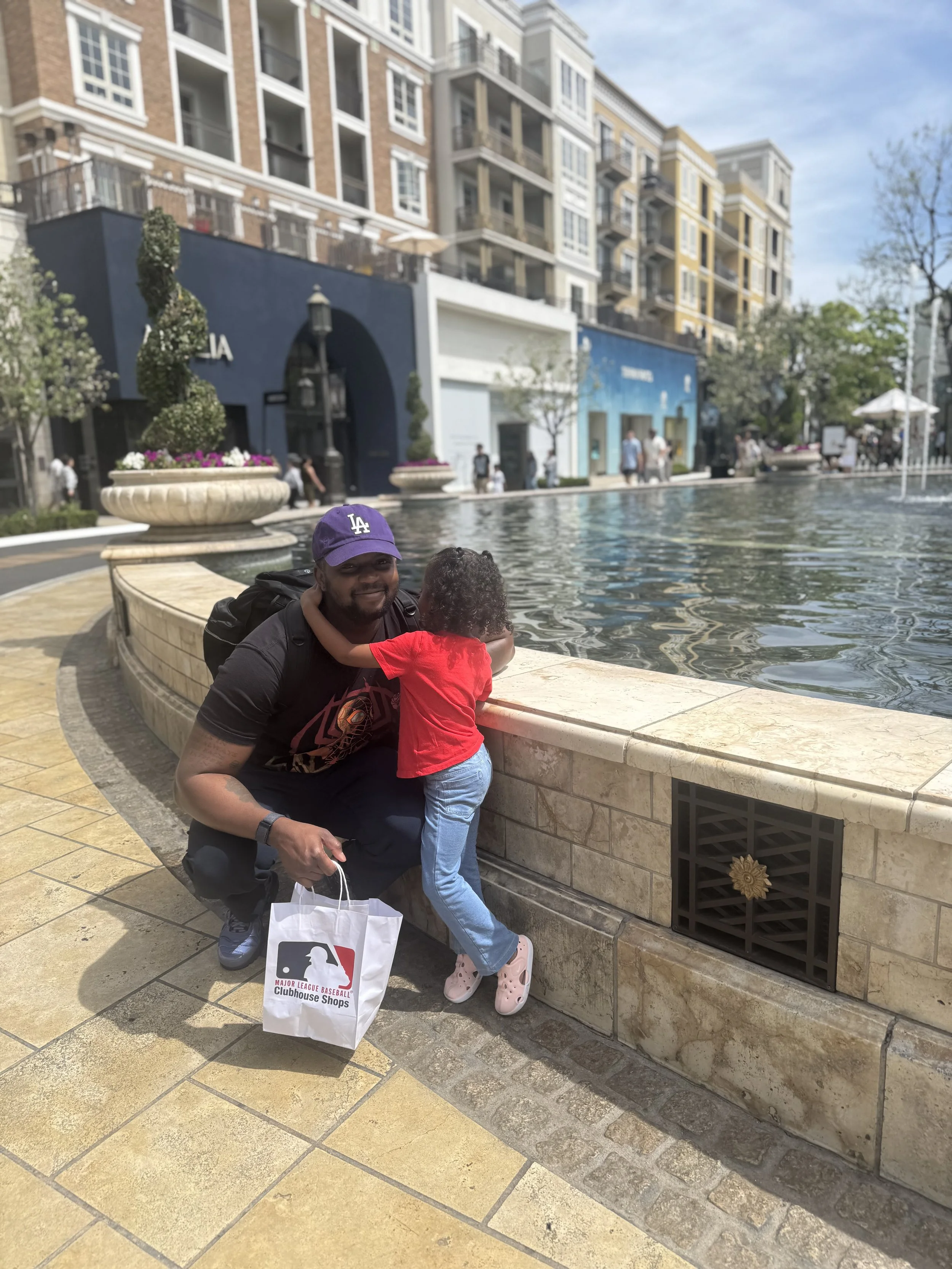 A man and a young girl are at a fountain in an urban area with tall apartment buildings. The man is smiling, wearing a purple 'LA' cap, gray shirt, black pants, and shoes. The girl is facing away, hugging the man, wearing a red shirt, blue jeans, and pink Crocs. They are beside a shopping bag and a baseball-themed shopping bag from Major League Baseball Clubhouse Shops.