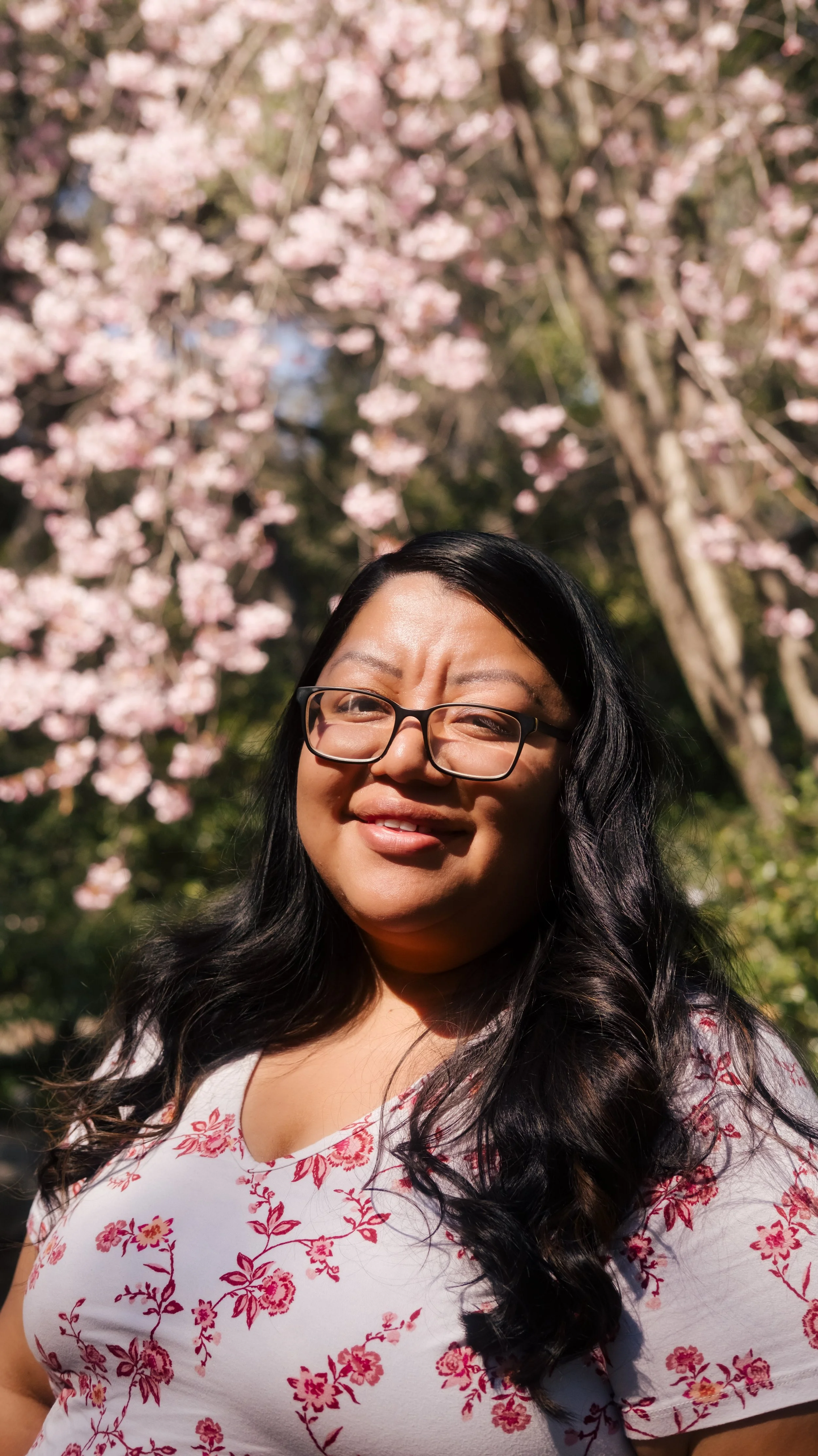 A woman with glasses and long dark hair smiling outdoors with pink cherry blossom trees in the background.