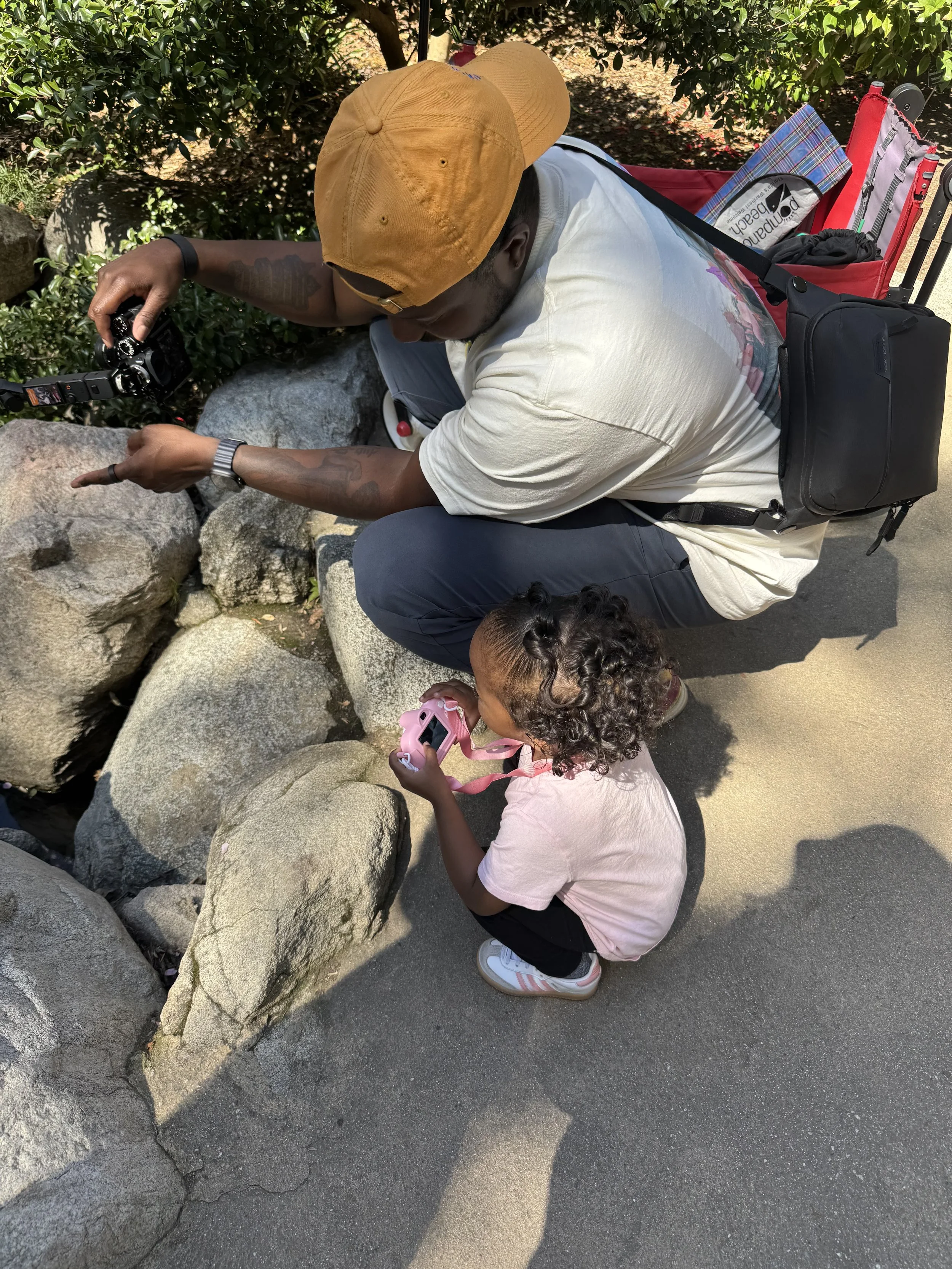 A man wearing a mustard yellow cap and white T-shirt squatting next to a young girl with curly hair and pink shoes, who is holding a pink camera, as they look at rocks or plants near a bush.