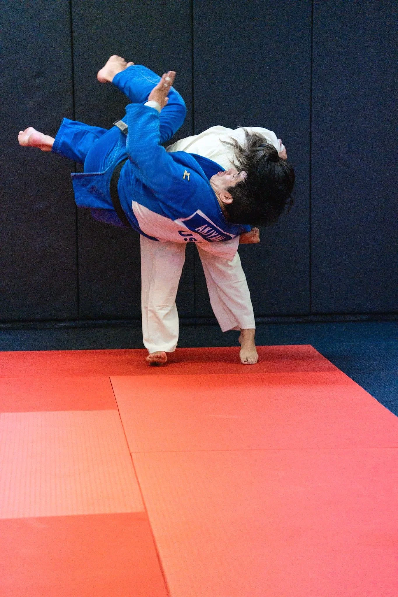 Two women practicing Brazilian Jiu-Jitsu on a red and black mat in a gym, with one woman wearing a blue gi and the other a white gi, grappling on the ground.
